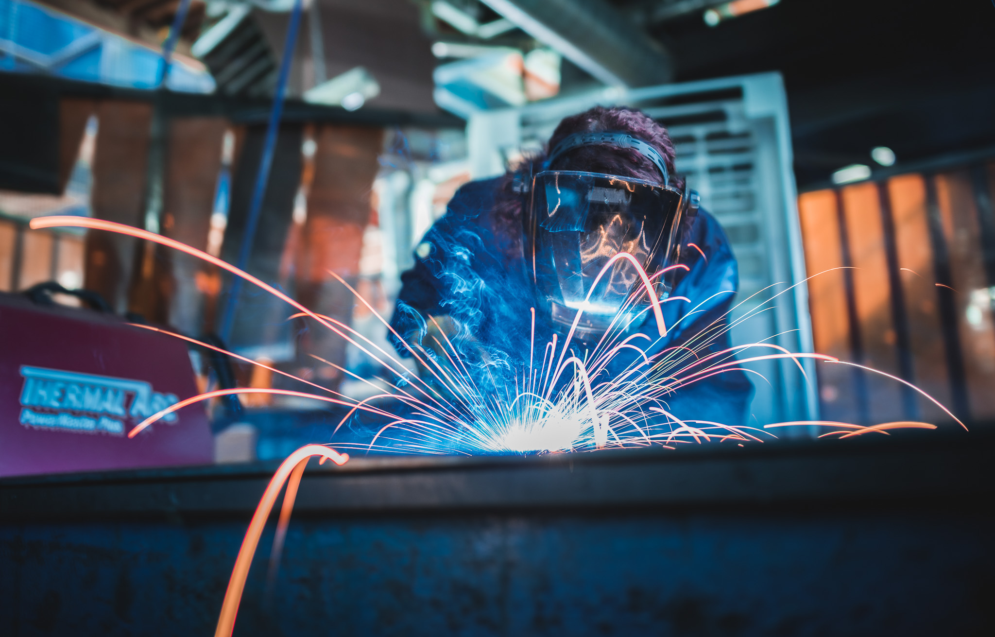 A person wearing protective gear and a face visor is welding metal, with bright sparks and blue light illuminating their work in an industrial setting.