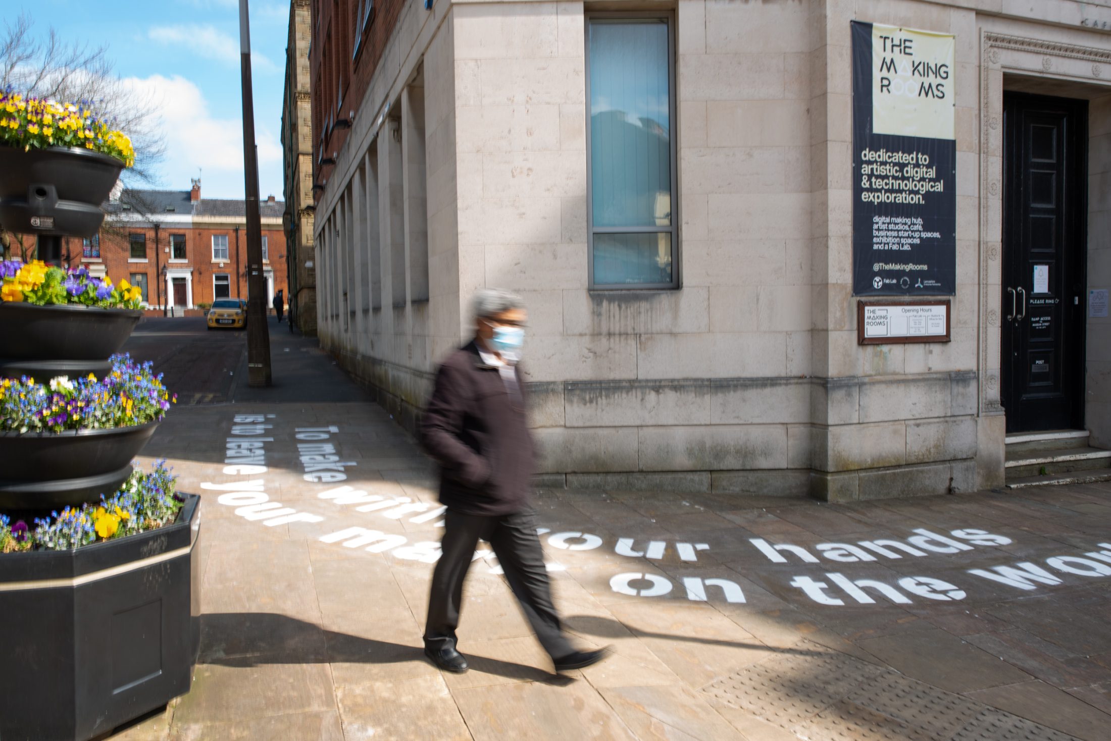A person wearing a mask walks past a building with a sign reading The Making Rooms. Poetry in white text is sprayed on the pavement and there are flower planters on the left side of the image.