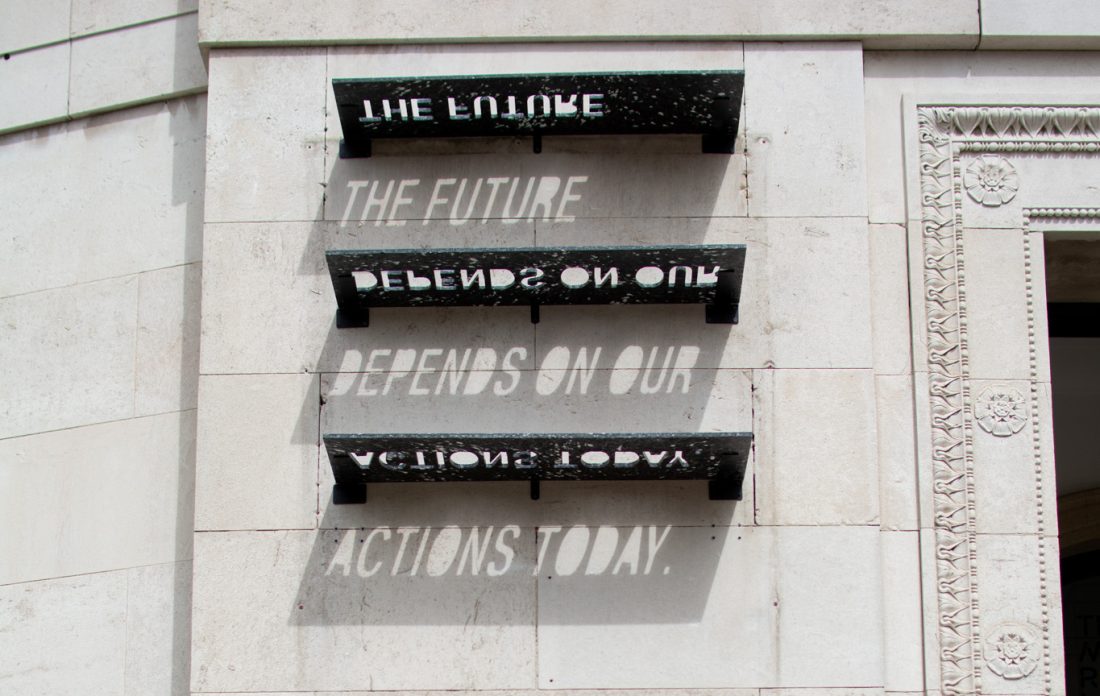Sculptural shelves on a white stone wall cast shadows that read: “THE FUTURE DEPENDS ON OUR ACTIONS TODAY.” The phrase forms only when sunlight passes through cut-out letters on the recycled plastic shelves.
