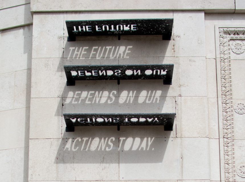 Sculptural shelves on a white stone wall cast shadows that read: “THE FUTURE DEPENDS ON OUR ACTIONS TODAY.” The phrase forms only when sunlight passes through cut-out letters on the recycled plastic shelves.