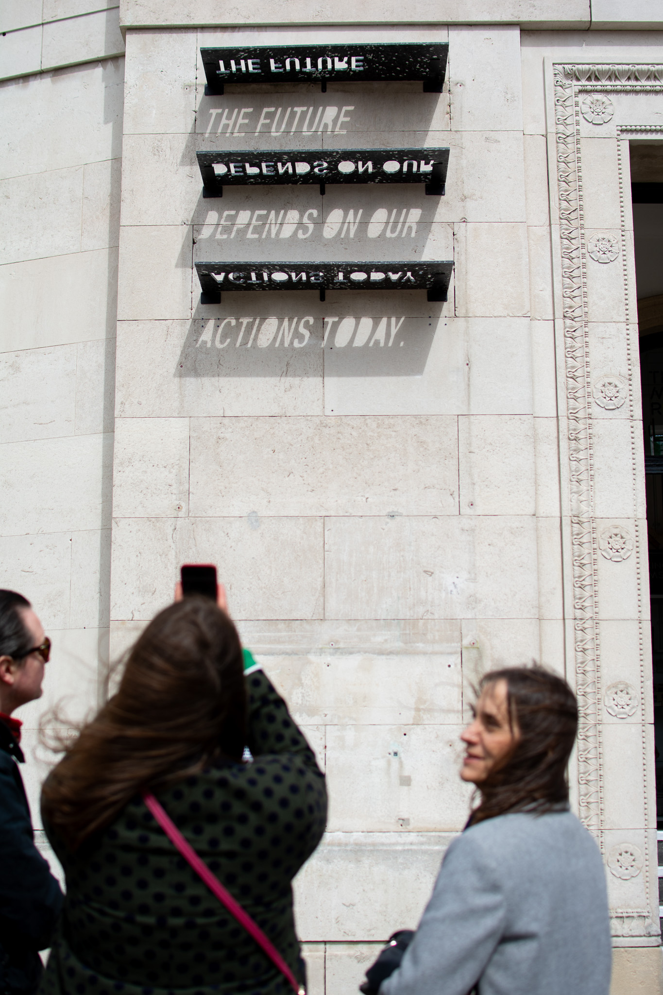 Three people look at an art installation on a building wall. Shadows from the installation spell out THE FUTURE DEPENDS ON OUR ACTIONS TODAY when sunlight hits it. One person takes a photo with a mobile.