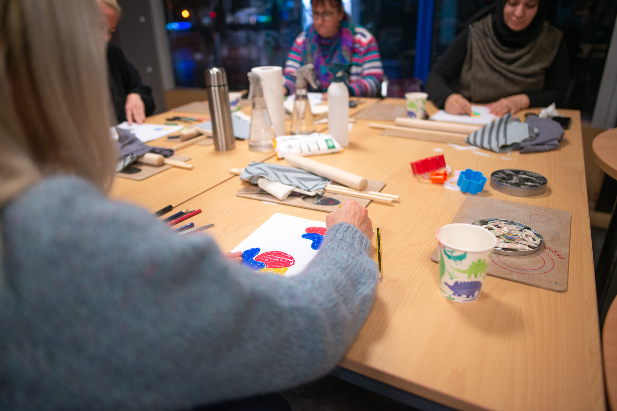 People gather around a wooden table, engaging in a workshop with coloured pencils and paper.
