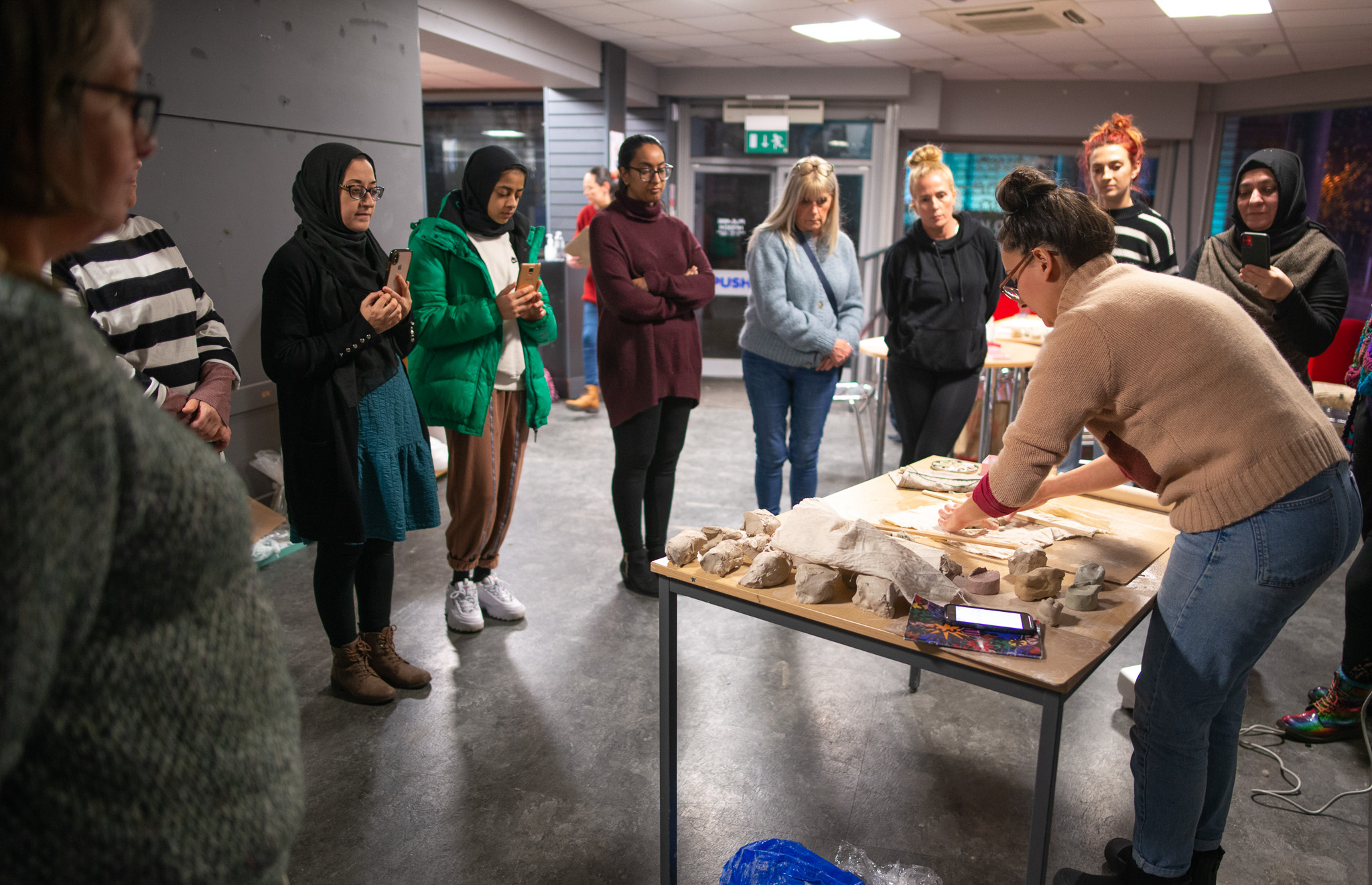 A group of people stands in a semicircle, attentively watching Aliyah Hussain demonstrating a hands-on activity at a table covered with art supplies and materials in a brightly lit indoor space.