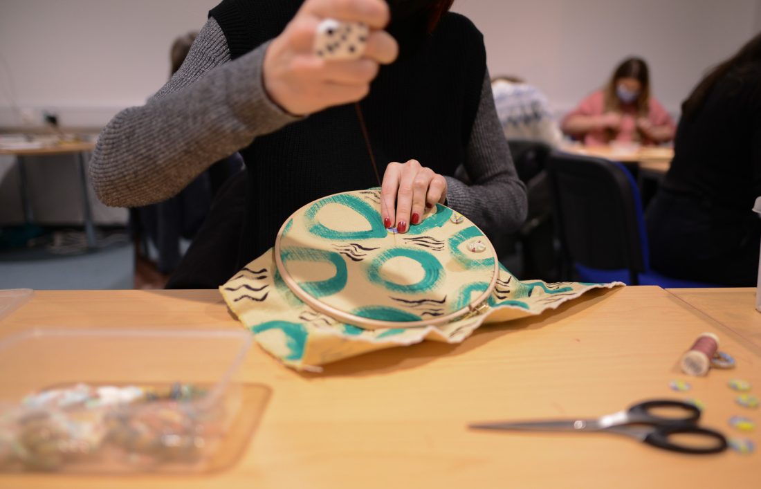 A person stitches a colourful, patterned fabric stretched over a circular frame at a table, with sewing tools and scissors nearby. Other people are working in the background.
