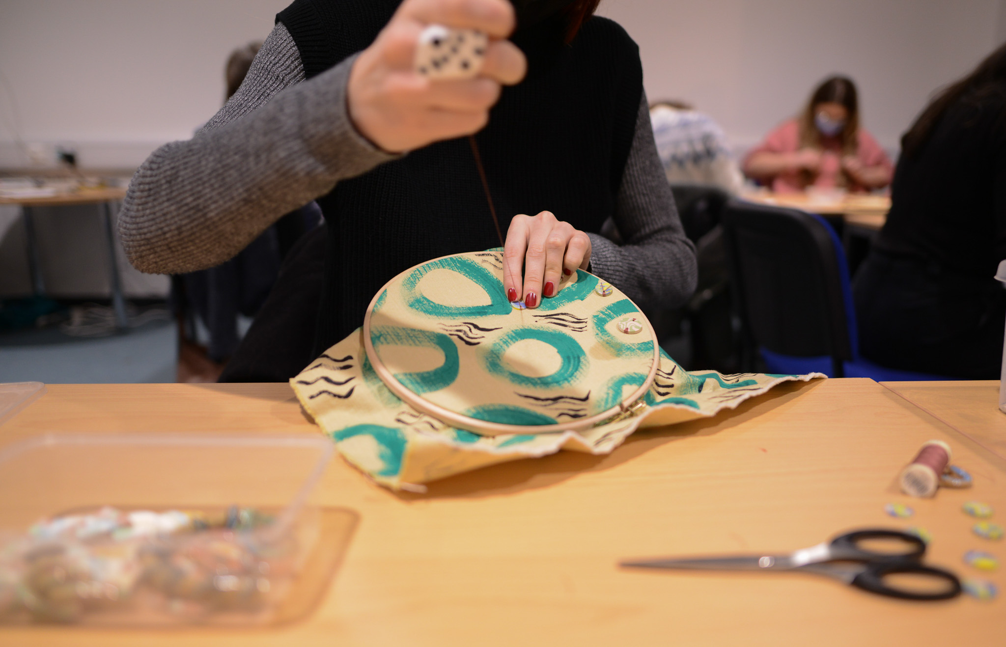 A person stitches a colourful, patterned fabric stretched over a circular frame at a table, with sewing tools and scissors nearby. Other people are working in the background.