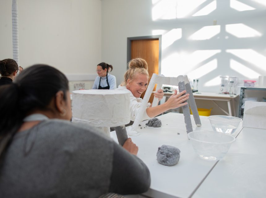People work on artworks paper and clay at a table in a bright studio. A woman in the centre smiles and reaches towards a sculpture, whilst others focus on their own work in the background.