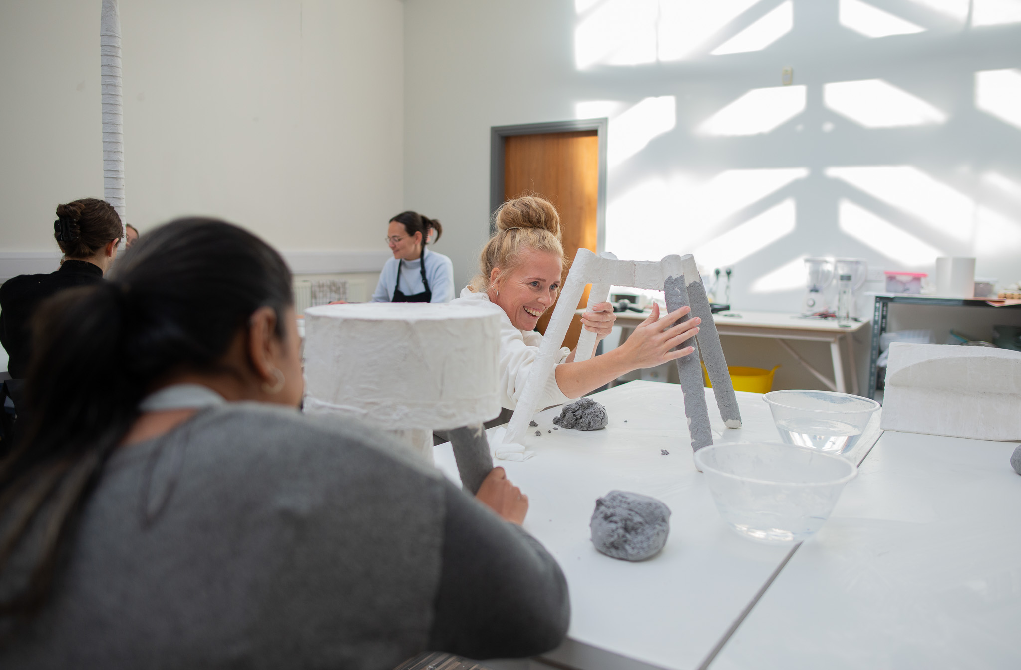 People work on artworks paper and clay at a table in a bright studio. A woman in the centre smiles and reaches towards a sculpture, whilst others focus on their own work in the background.