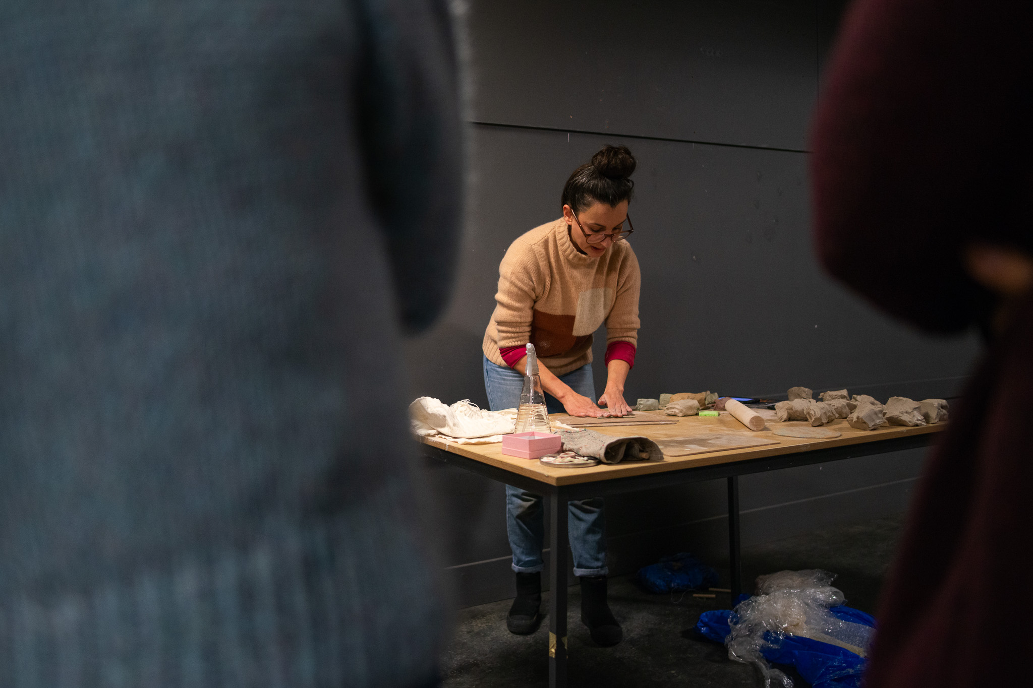 Aliyah Hussain works with clay on a table, surrounded by art materials, in a dimly lit room with people observing in the foreground.