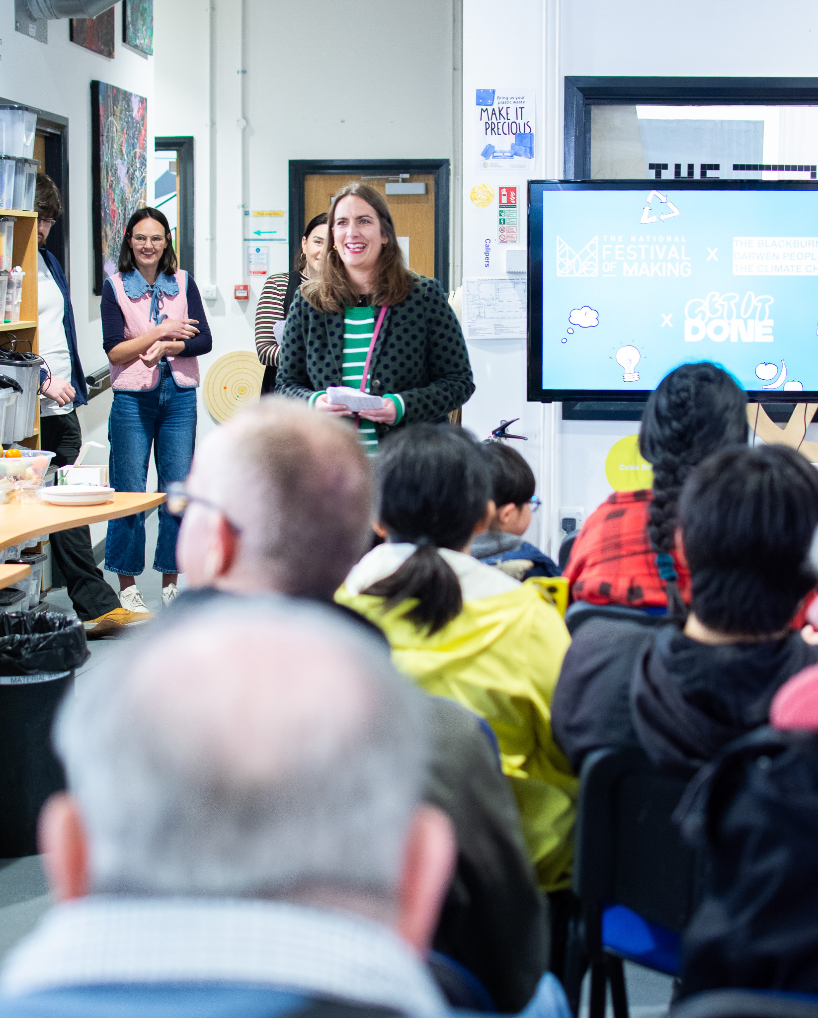 A woman speaks at an event, engaging an attentive audience. A screen displays event details in the background.