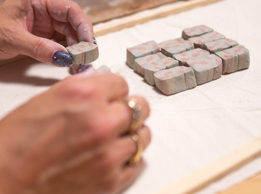 A person with painted nails arranges small, rectangular clay pieces with a marbled pattern on a fabric-covered tray. A rolling pin and other clay tools are visible in the background.