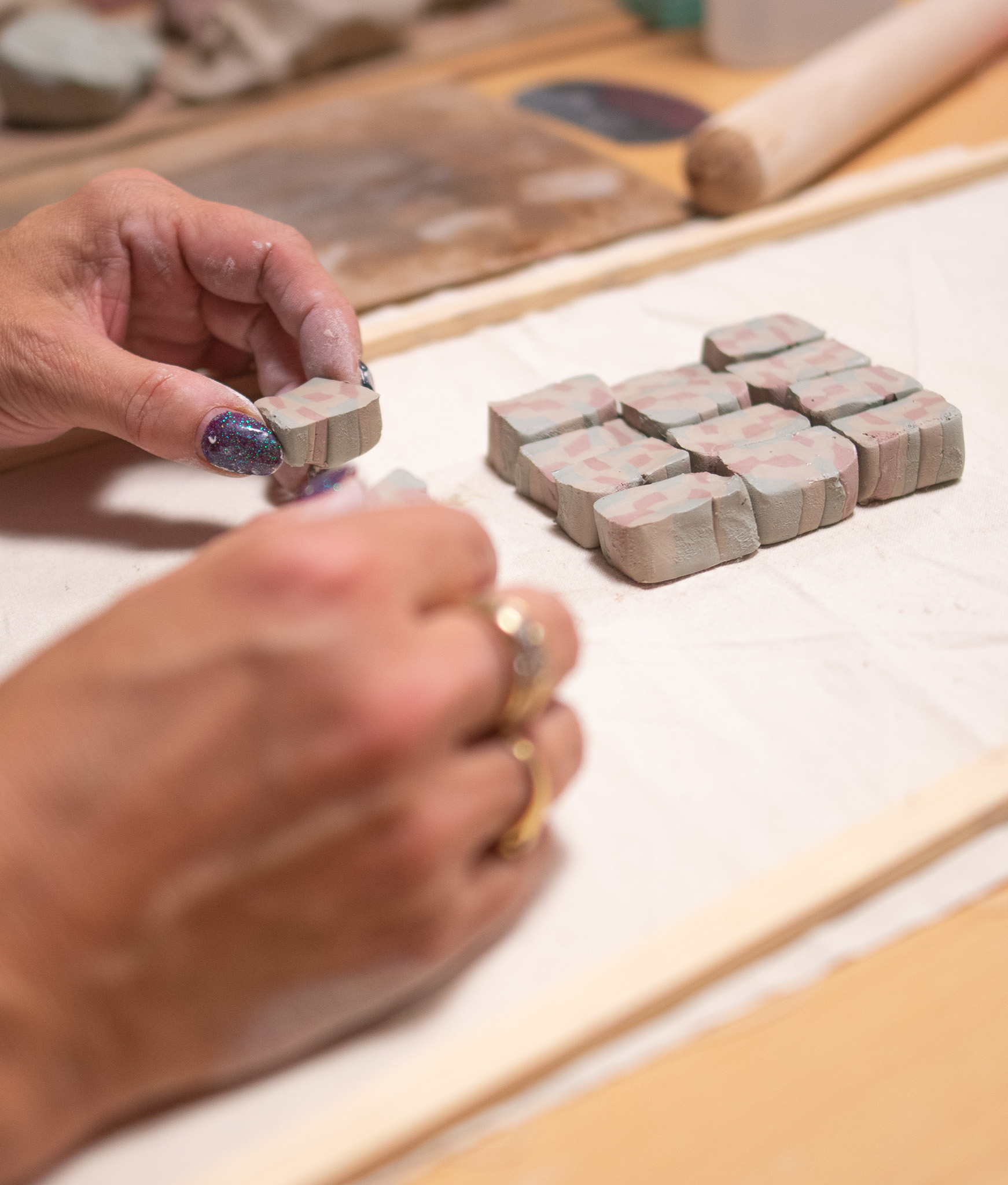 A person with painted nails arranges small, rectangular clay pieces with a marbled pattern on a fabric-covered tray. A rolling pin and other clay tools are visible in the background.