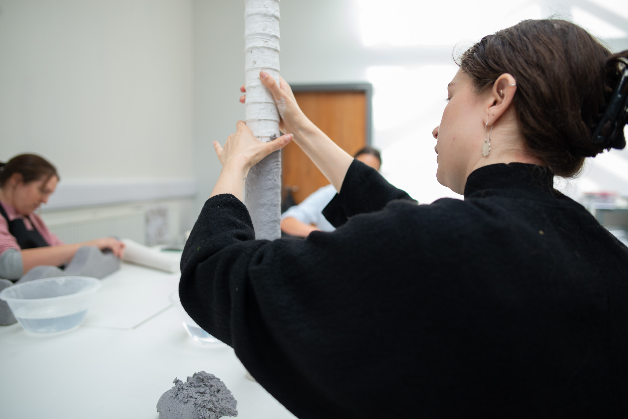 A woman in a black sweater molds a tall, cylindrical clay sculpture in a bright room. Another person, focused, works in the background.