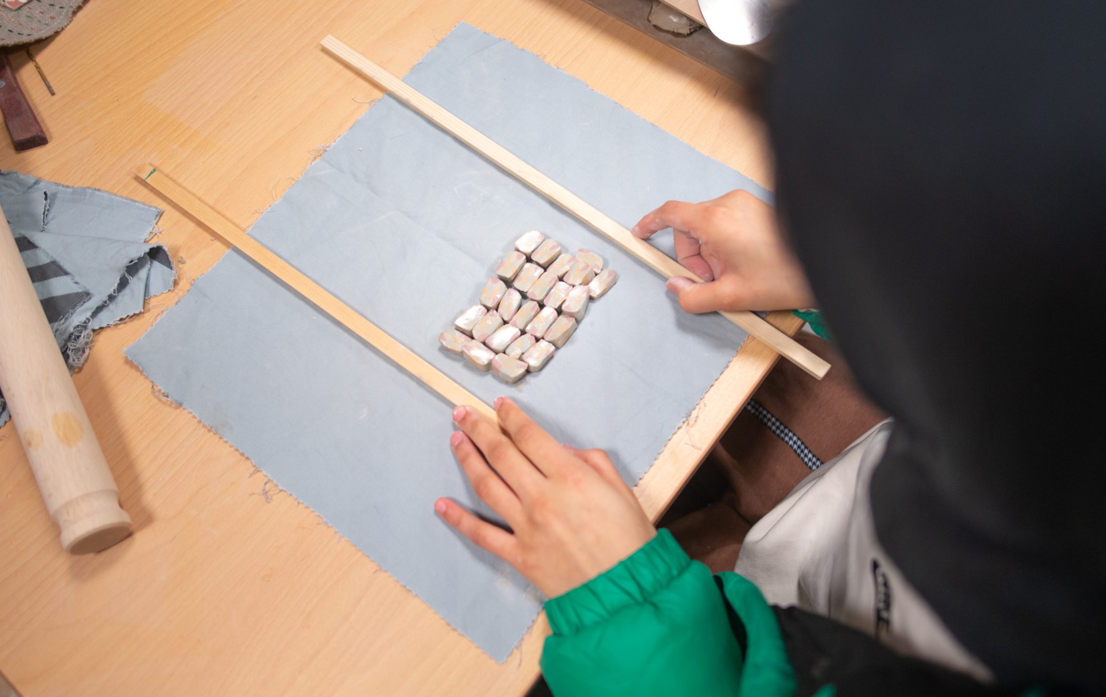 A person in a green jacket uses two wooden sticks to measure and align small rectangular pieces of clay on a blue cloth placed on a wooden table.