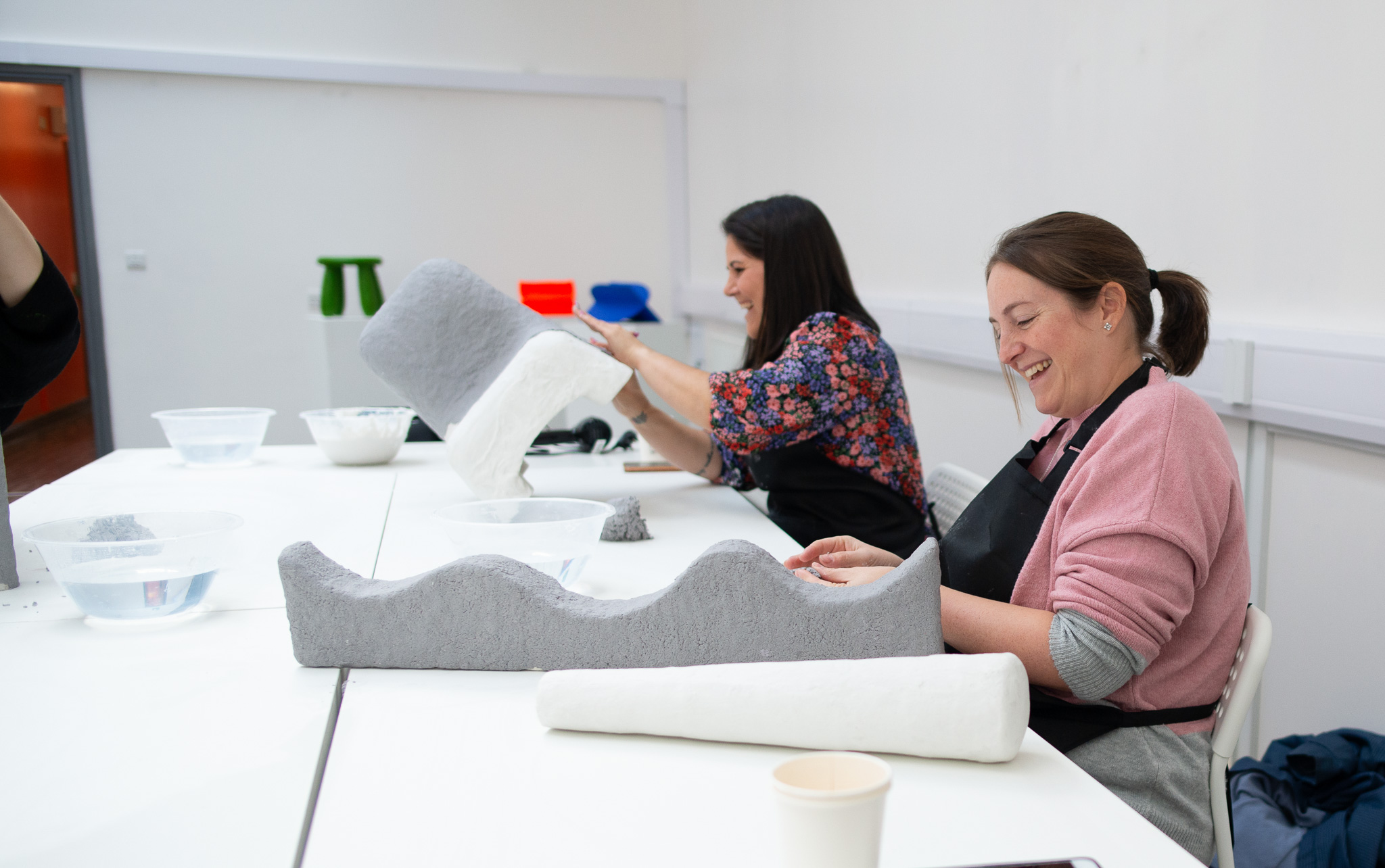 Two women in a bright room smile while sitting at a table in a workshop, working with paper clay and bowls of water.