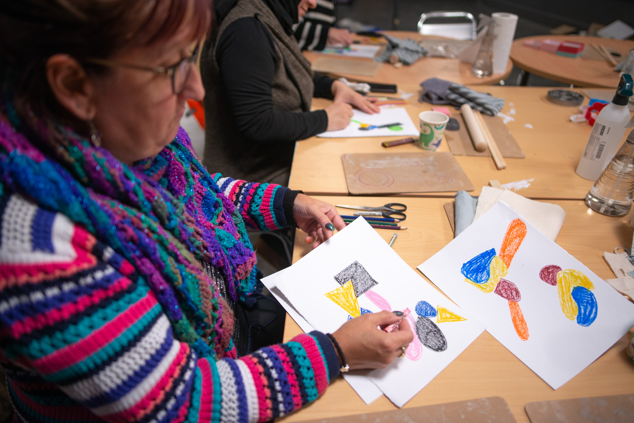A person in a striped crocheted jumper draws a colourful abstract picture on white paper at a wooden table, surrounded by art supplies.