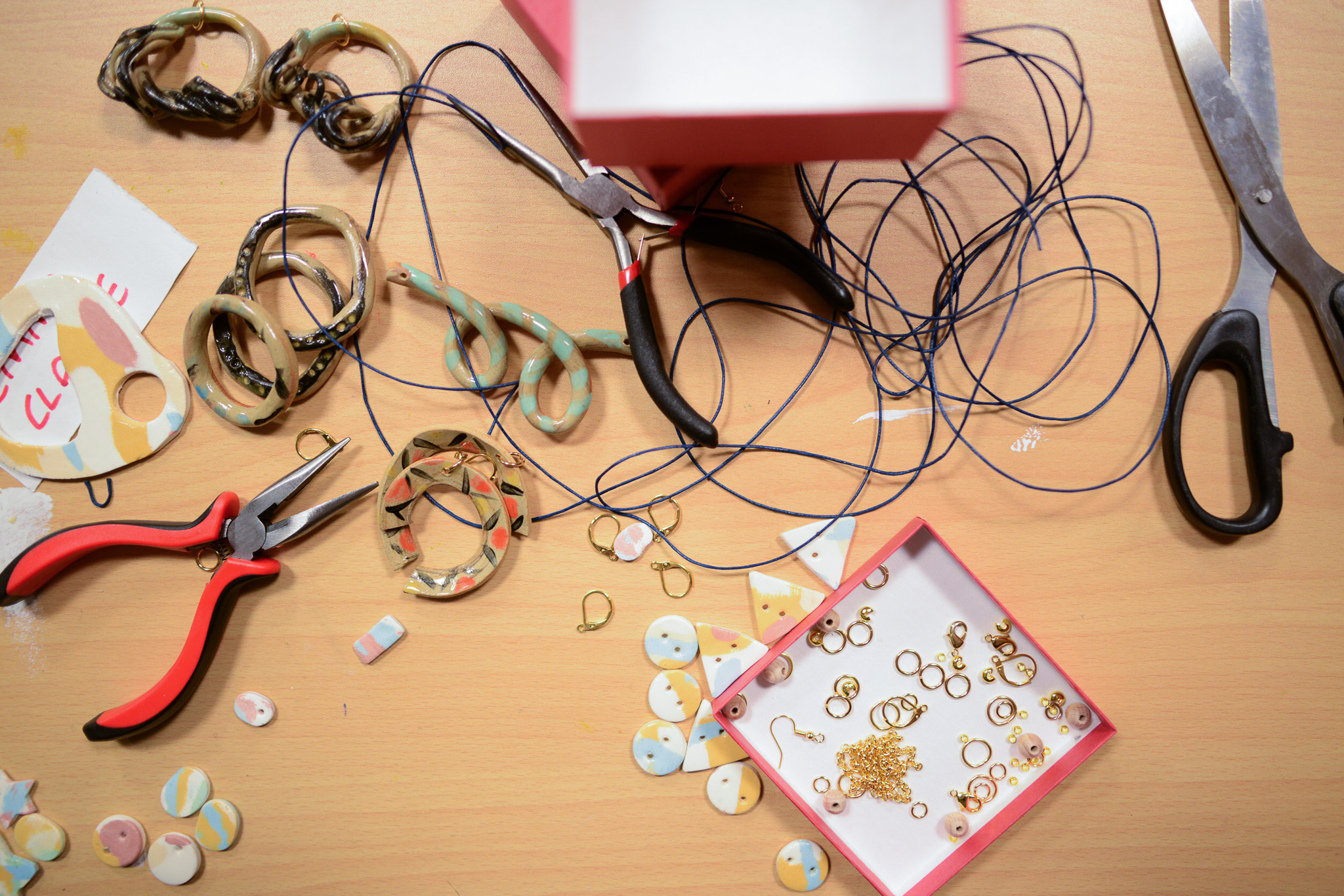 A tabletop with jewellery-making tools, bracelets, scissors, pliers, blue string, and a pink box containing gold rings and small charms, alongside colourful round charms and unfinished jewellery pieces.