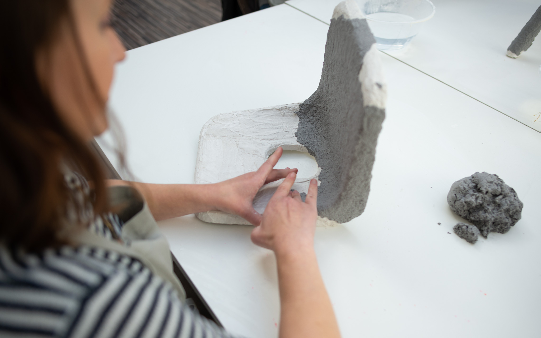A person adds paper clay to a white chair on a table, with a bowl of water and a lump of clay nearby.
