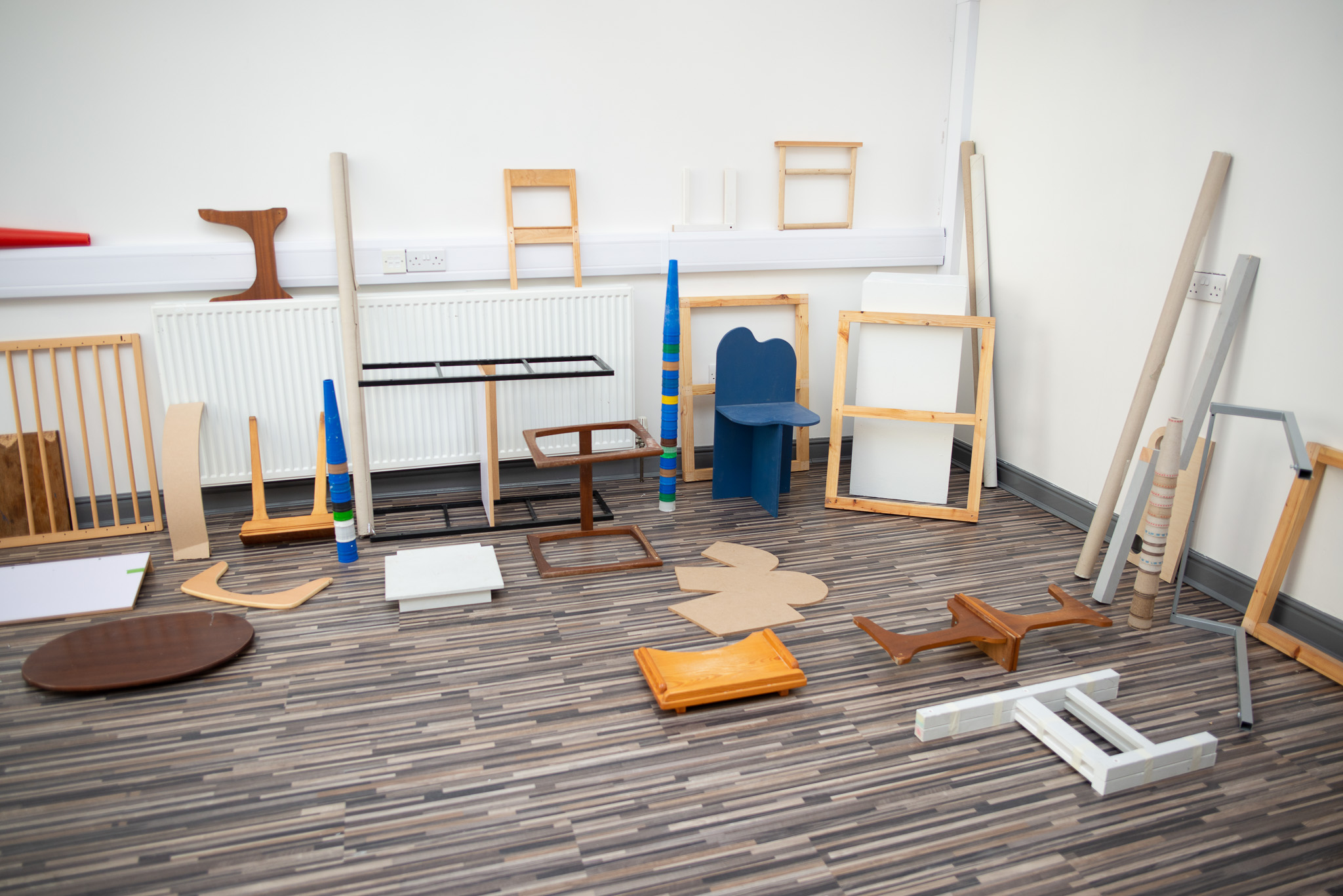 A variety of wooden pieces, frames and furniture are arranged on a striped floor near a white wall.