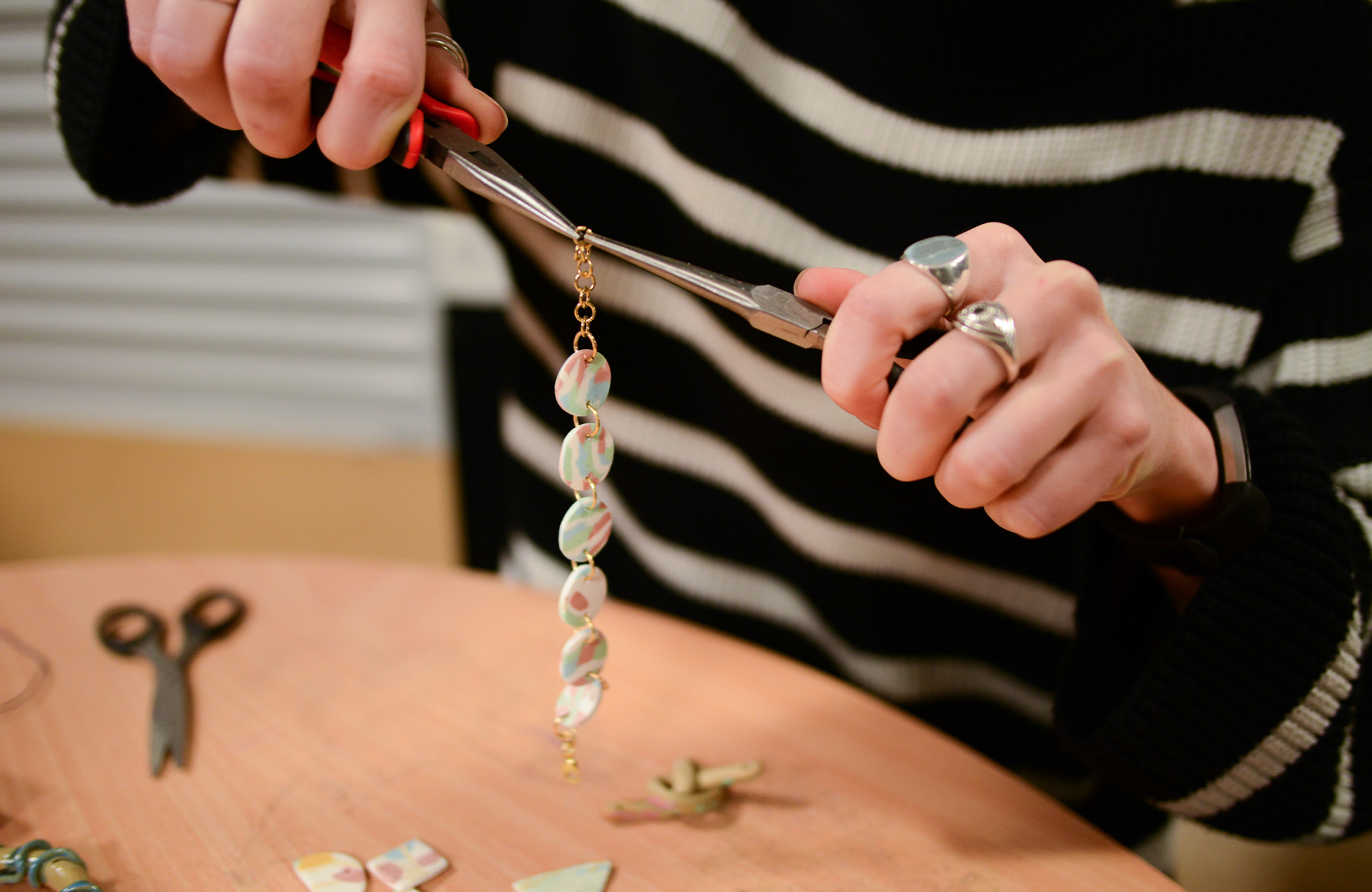 A person wearing a black and white striped jumper uses pliers to assemble a hanging jewellery piece made of ceramic beads at a table.