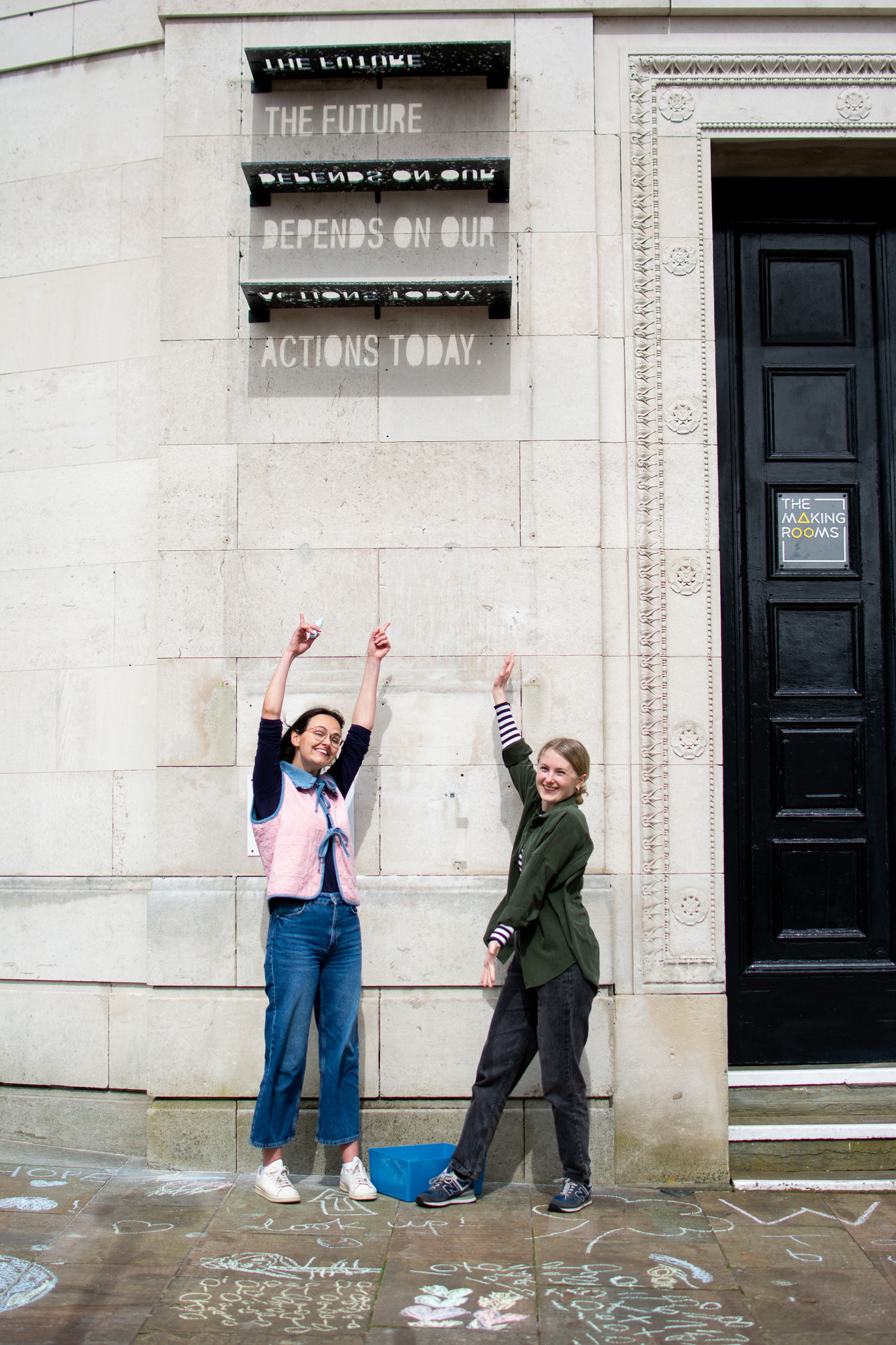 Two people stand outside a stone building, smiling and pointing up at a sign that reads THE FUTURE DEPENDS ON OUR ACTIONS TODAY. Chalk drawings are on the pavement near their feet.
