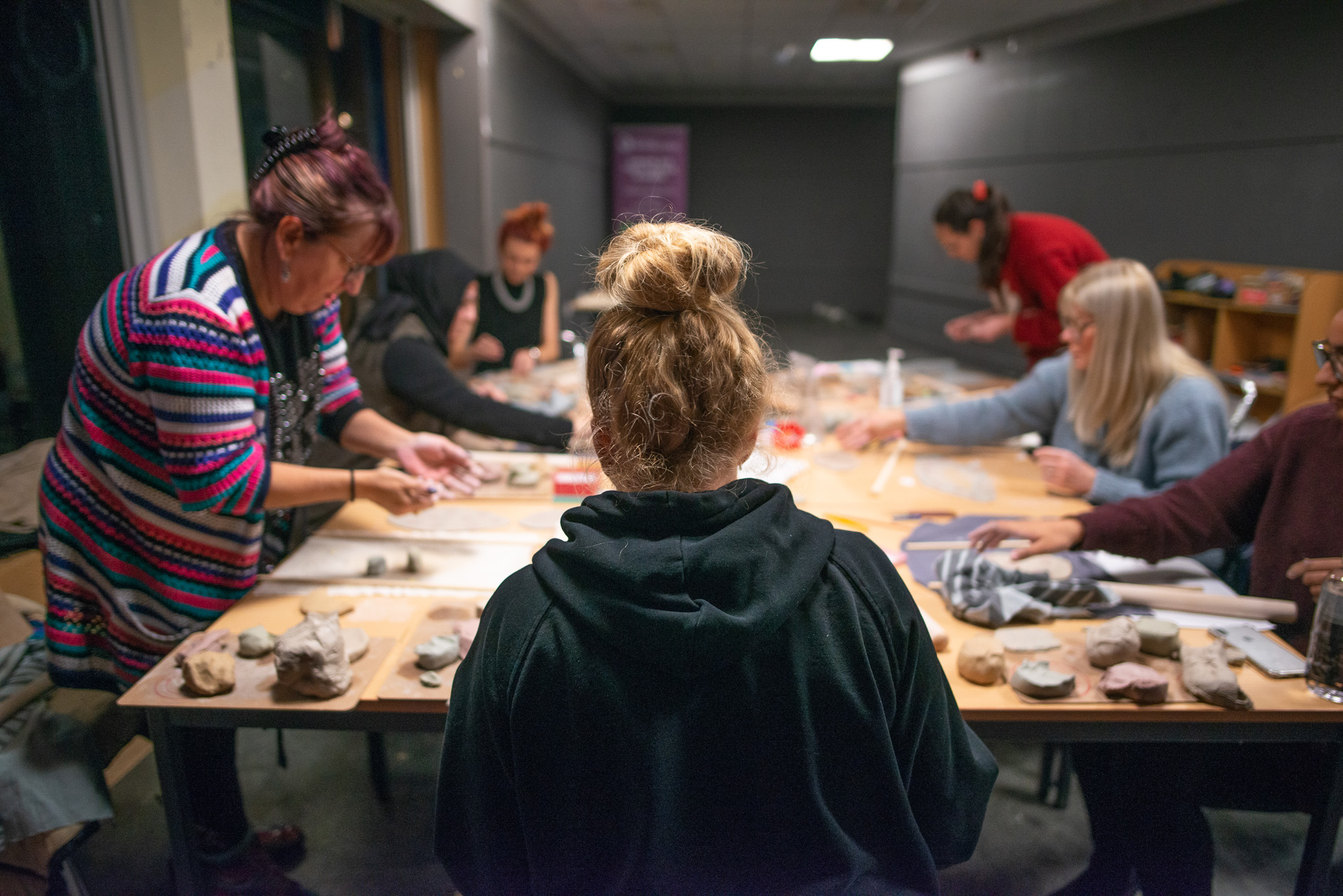 A group of people are seated around a table, focused on making with clay and various tools in a well-lit room. One person in a black hoodie is seated in the foreground, facing the table.
