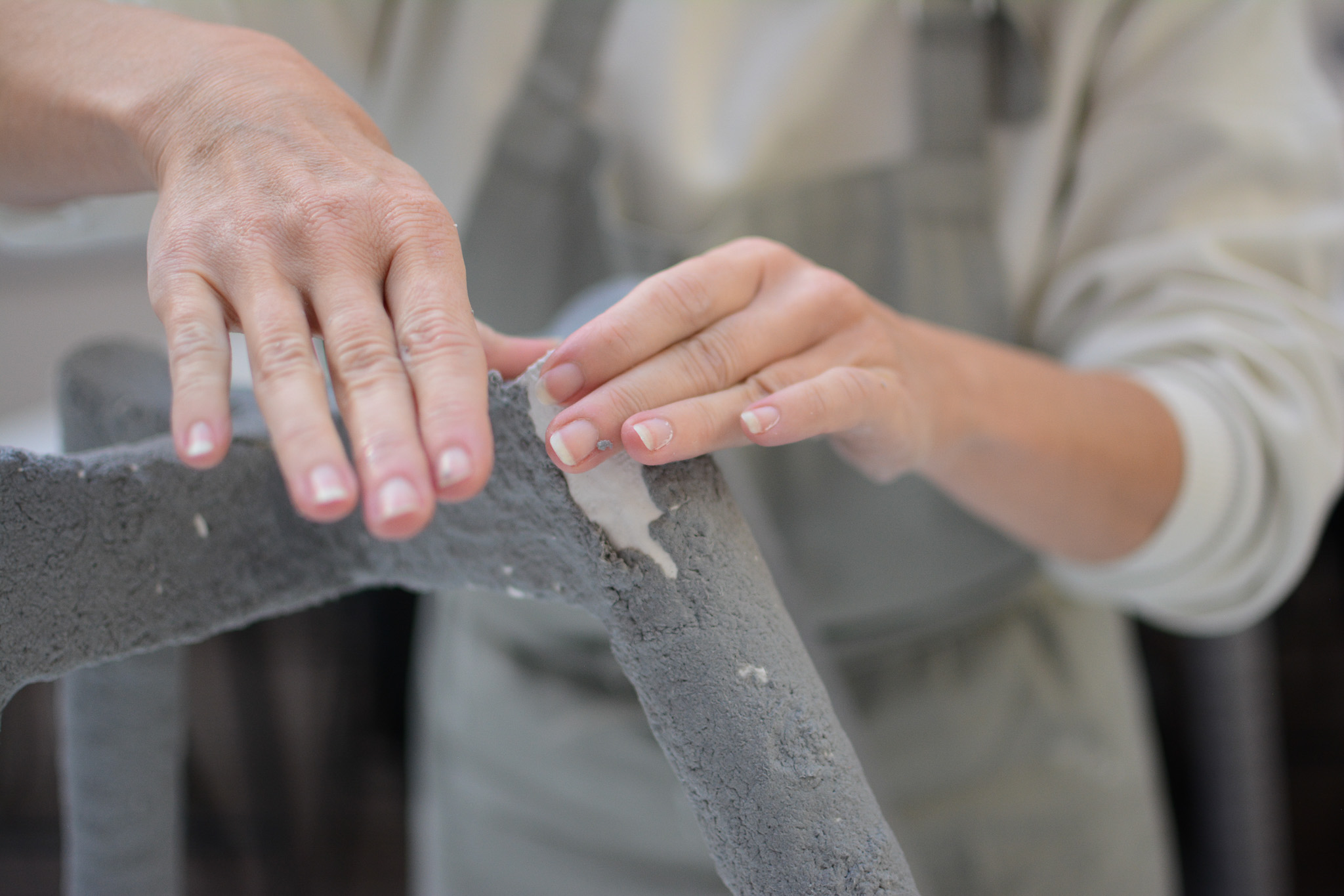 Close-up of a person’s hands smoothing over a rough, grey, textured surface with paper clay, wearing a light-coloured long-sleeve shirt and apron.