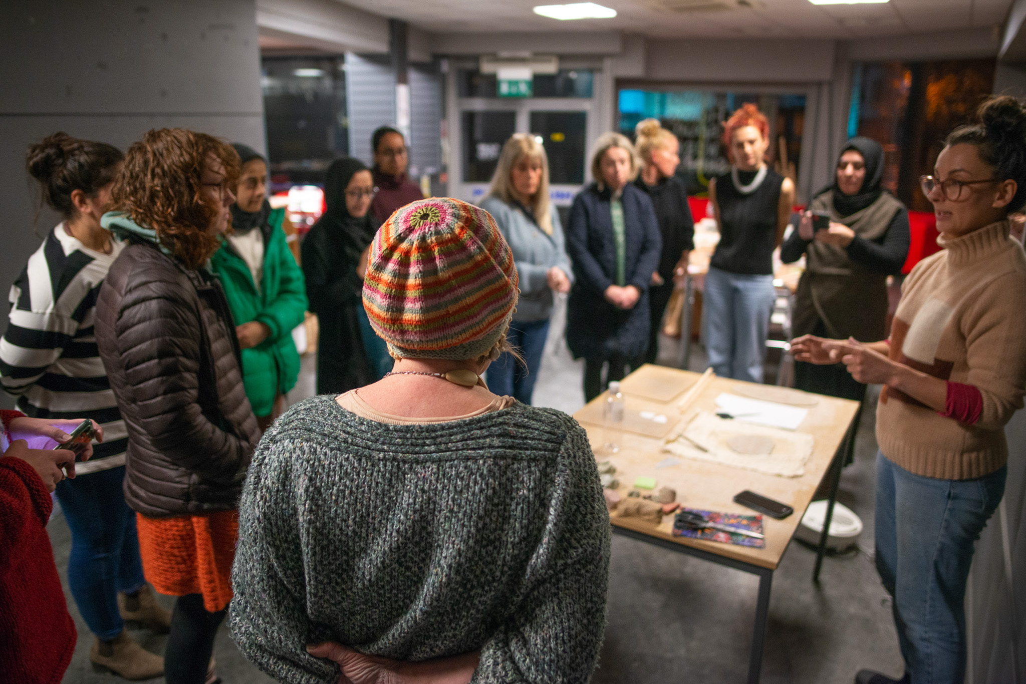 A group of women stand in a circle indoors, listening to the artist speaking and gesturing near a table with art supplies.