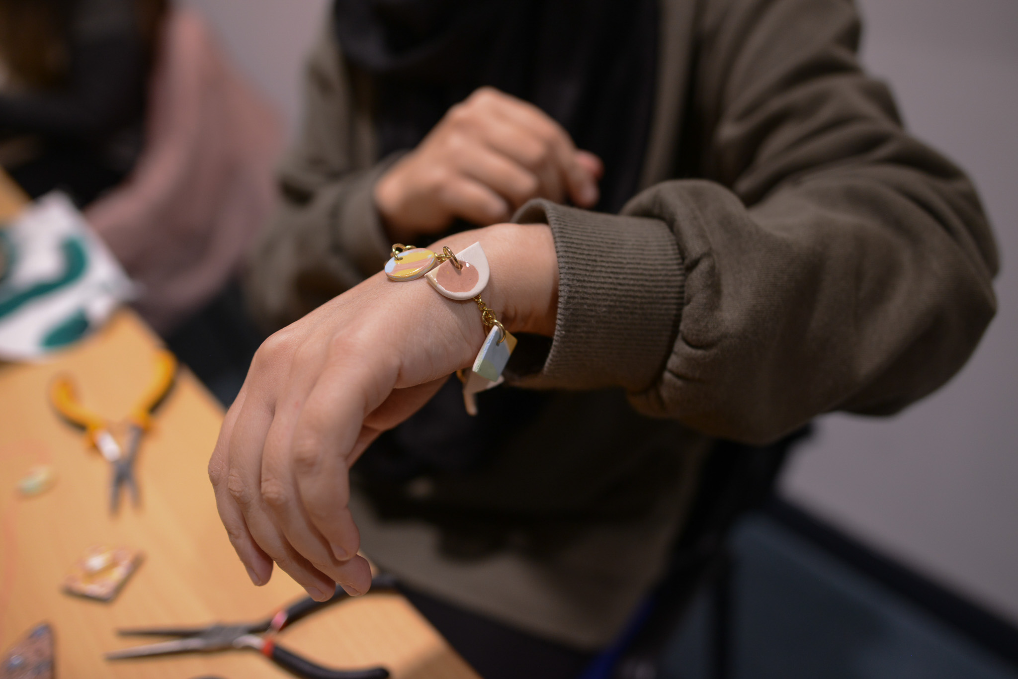 A person wearing a greenish-brown jumper displays a handmade bracelet with pastel-coloured ceramic charms. Crafting tools and materials are on the wooden table nearby.