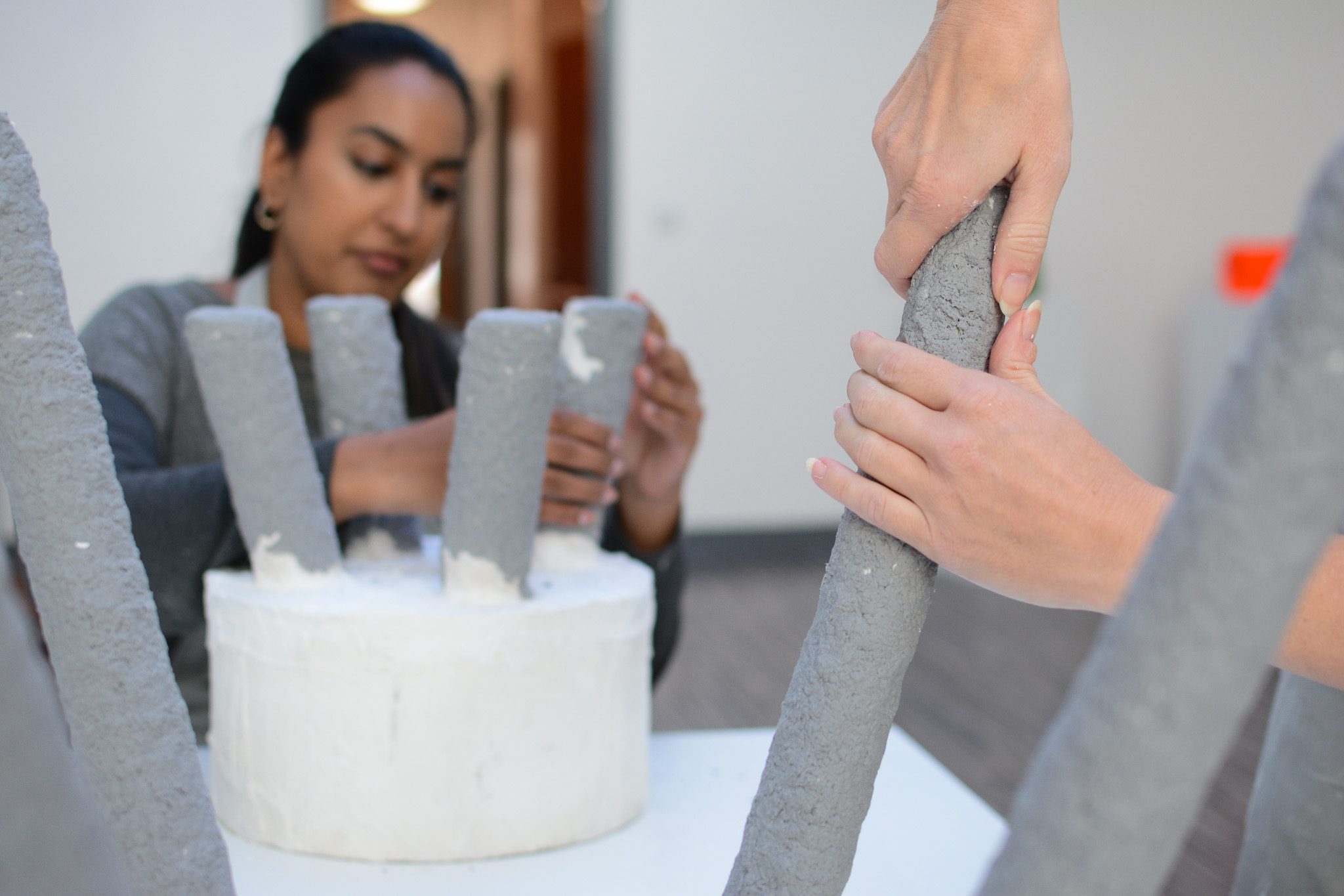 Two people work together on a grey textured sculpture made of cylindrical forms in a bright room.
