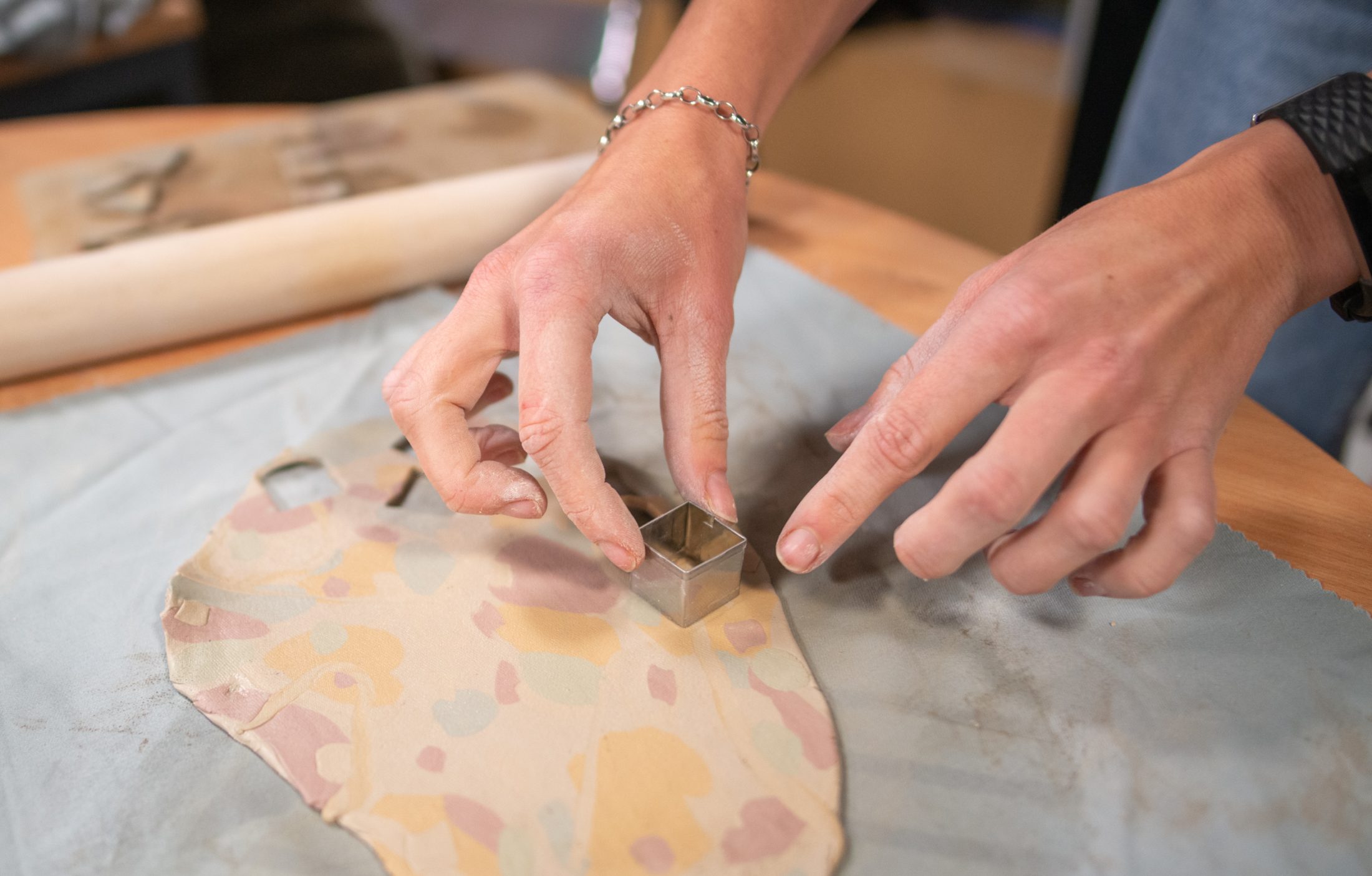 Close-up of hands using a small metal square cutter on rolled-out, multicoloured clay, with a rolling pin and tools on the table in the background.