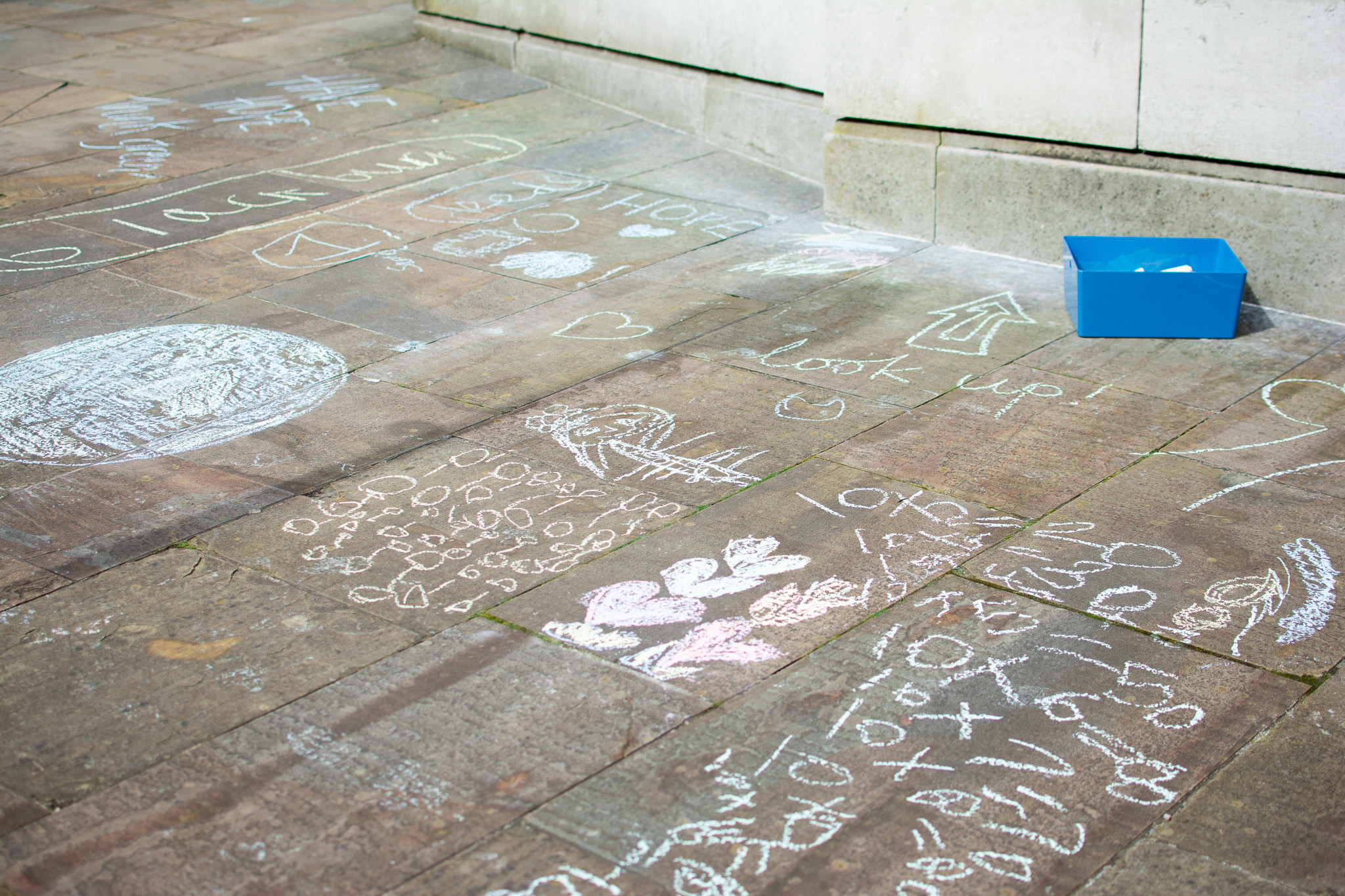 Colourful chalk drawings and writings cover a stone pavement, including shapes, hearts, and the words look up. A blue container with chalk pieces sits in the top right corner near a stone wall.