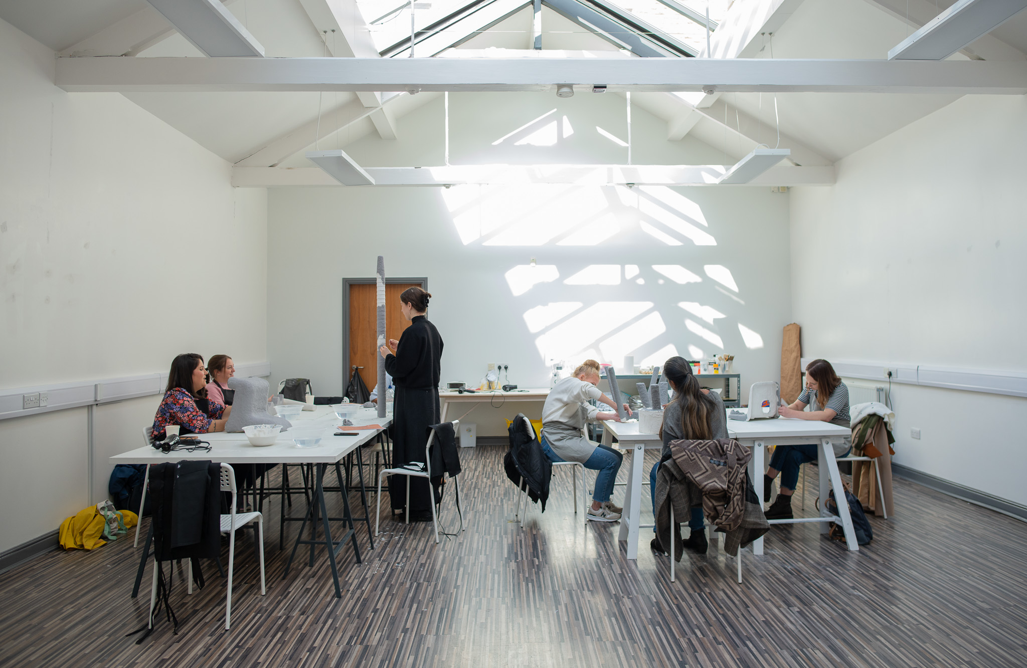 A group of people work and talk at tables in a bright, room with high ceilings and skylights. Sunlight streams in, creating patterns on the white walls and wood-patterned floor.