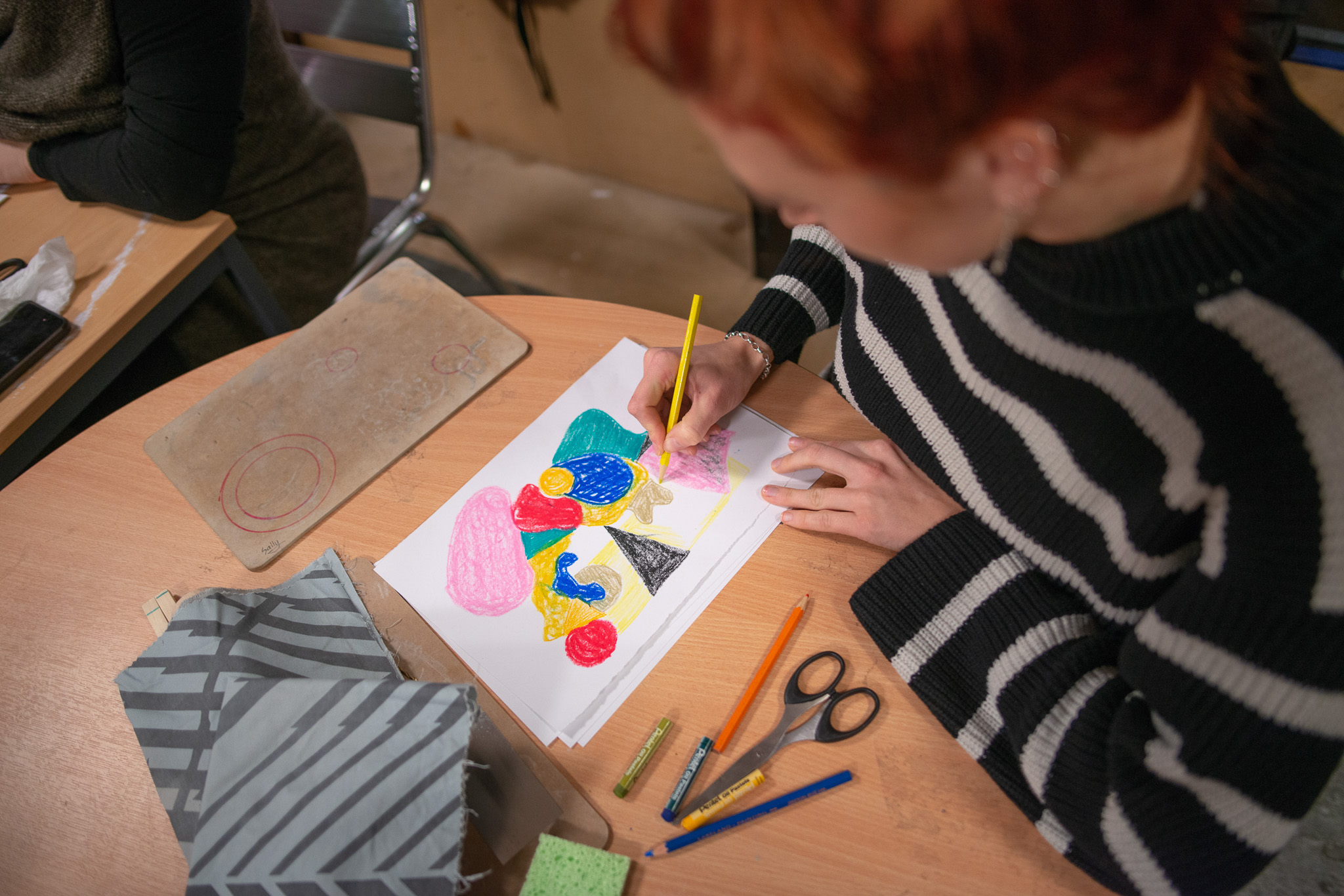 A person with red hair and a striped jumper draws a colourful abstract picture on white paper at a wooden table, surrounded by art supplies like felt-tip pens, pencils, and scissors.
