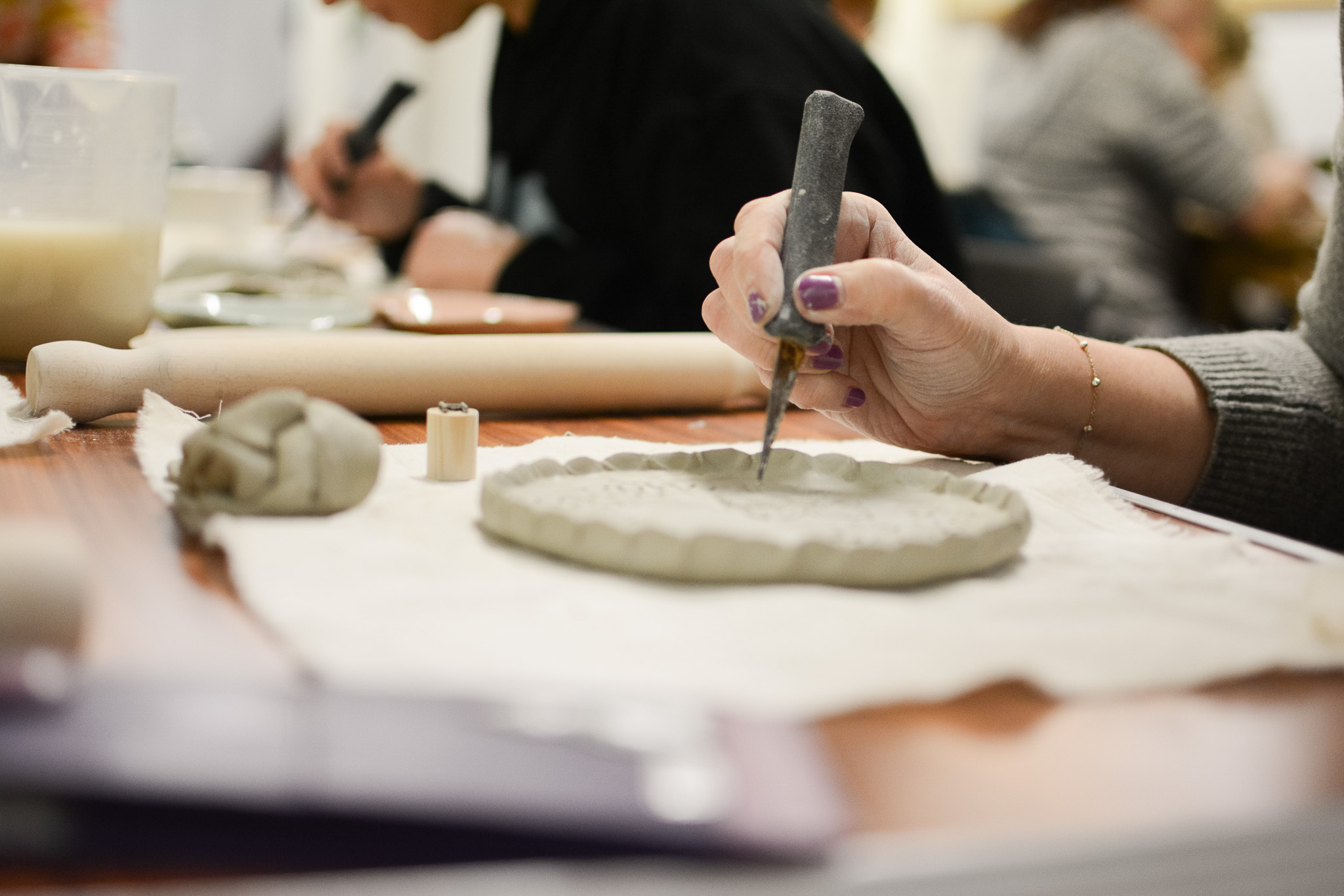 A person uses a carving tool to shape a piece of clay on a table, surrounded by other pottery tools and supplies, whilst others work in the background.