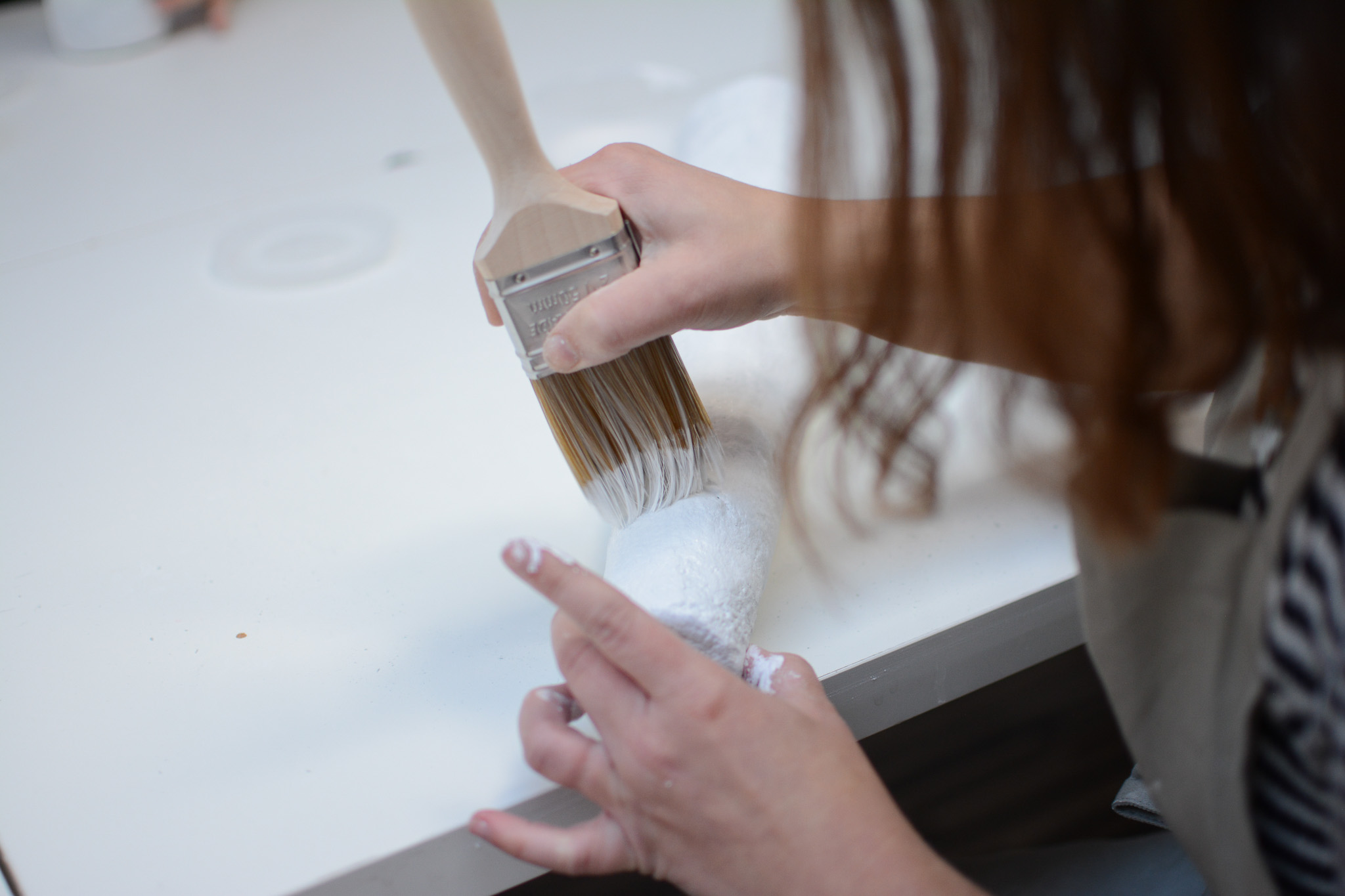 A person uses a large paintbrush to apply white paint to an object on a table.