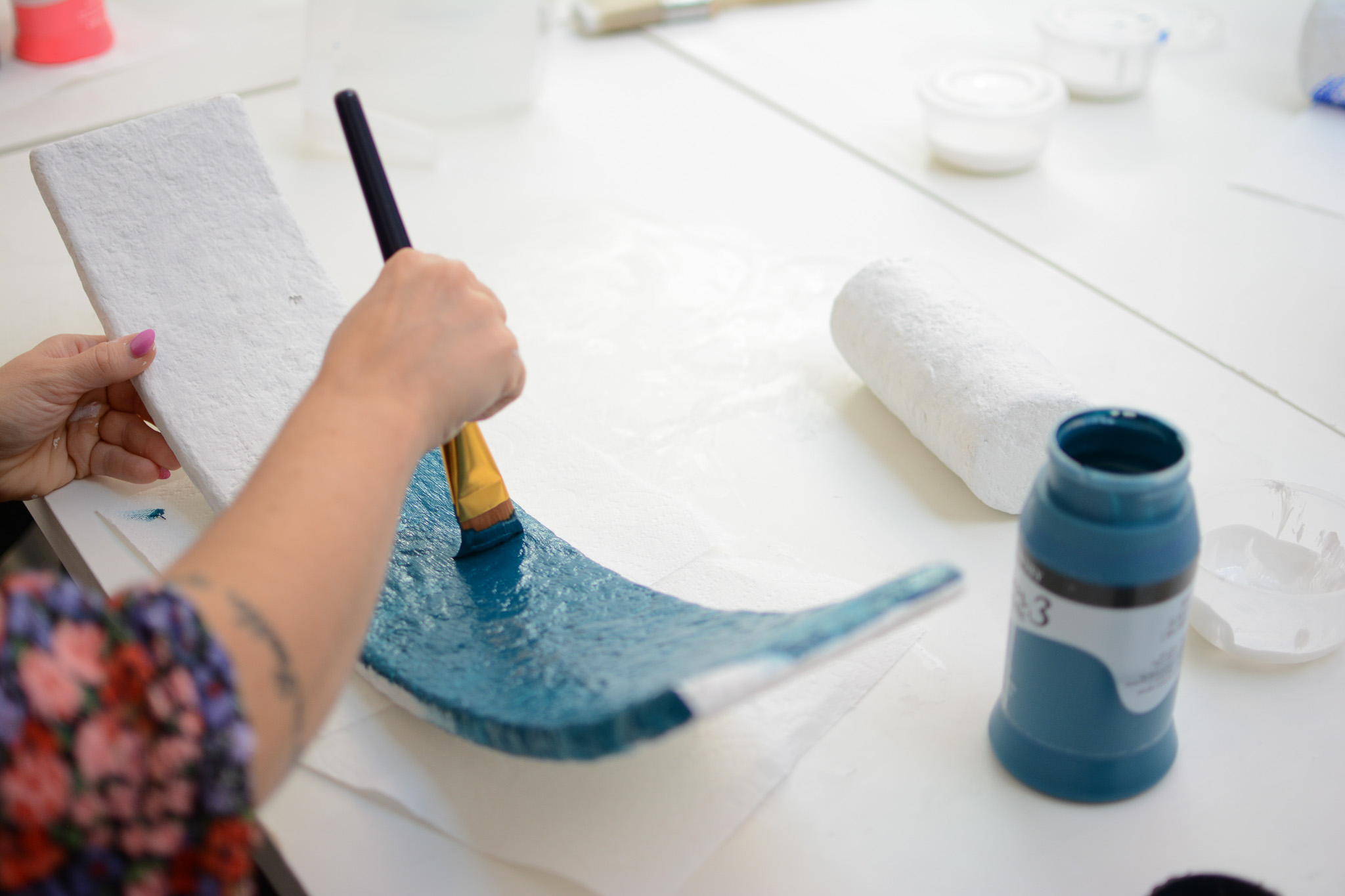A person paints a curved white object with blue paint, holding it in one hand, with painting supplies spread across a white table.