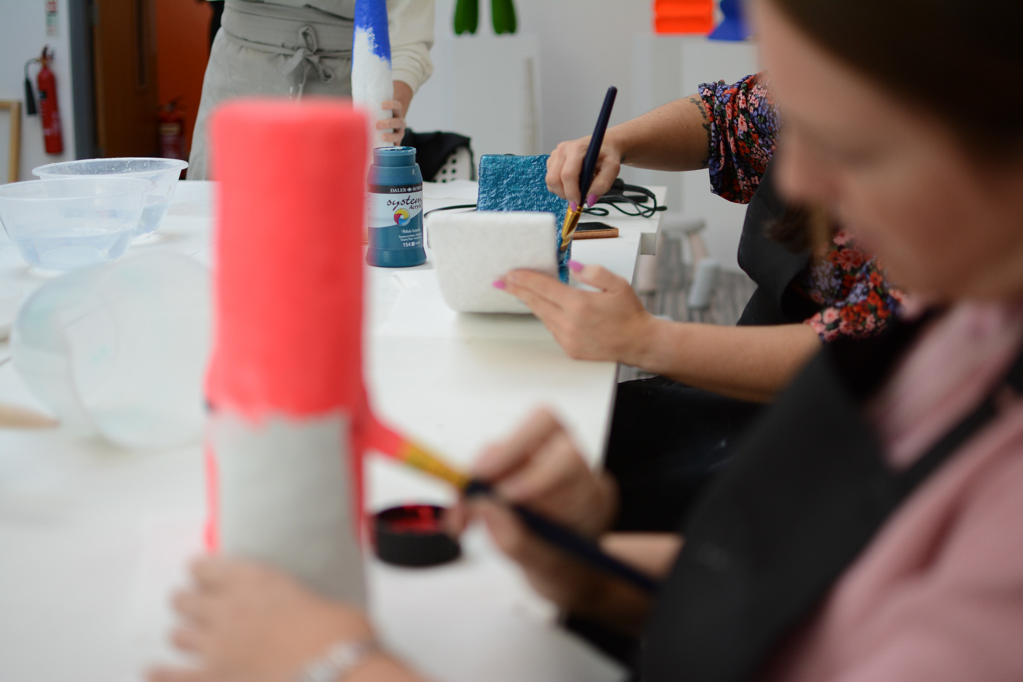 Close-up of people painting cylindrical and rectangular sculptures in bright colours like pink and blue at a workshop.