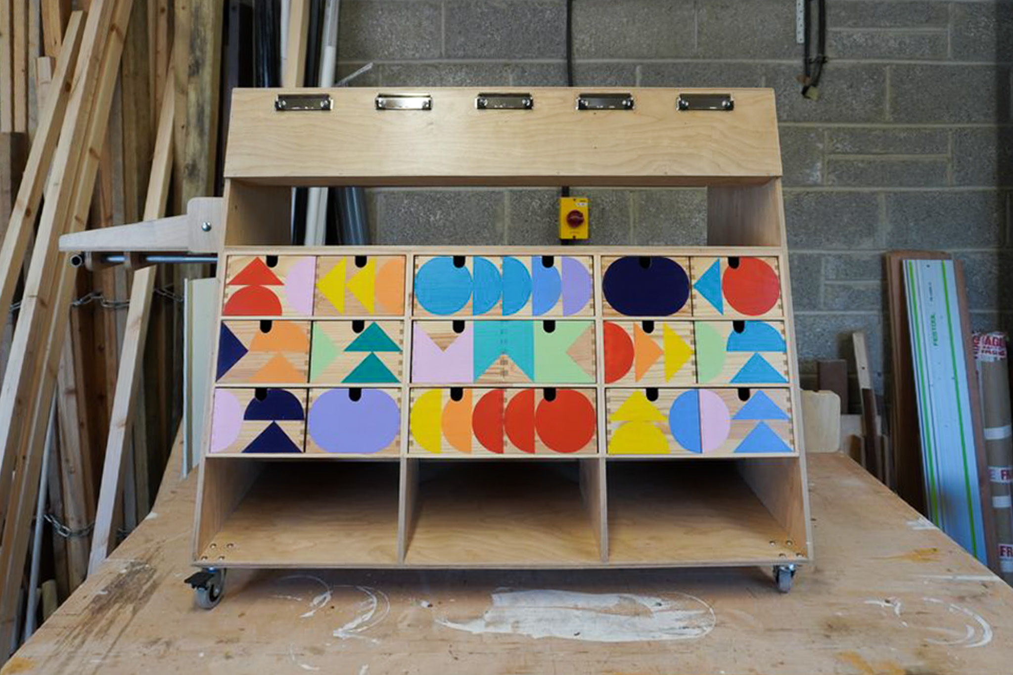A wooden cupboard with colourful, geometric-patterned drawer fronts stands on castors in a workshop with wooden planks and concrete walls in the background.