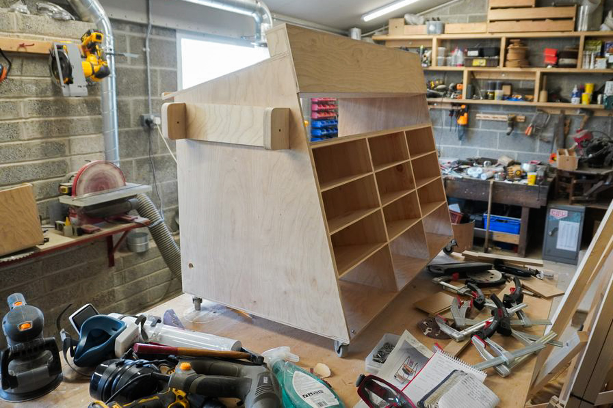 A wooden cabinet on wheels sits on a workbench in a busy workshop, surrounded by tools, clamps, and shelves of equipment.
