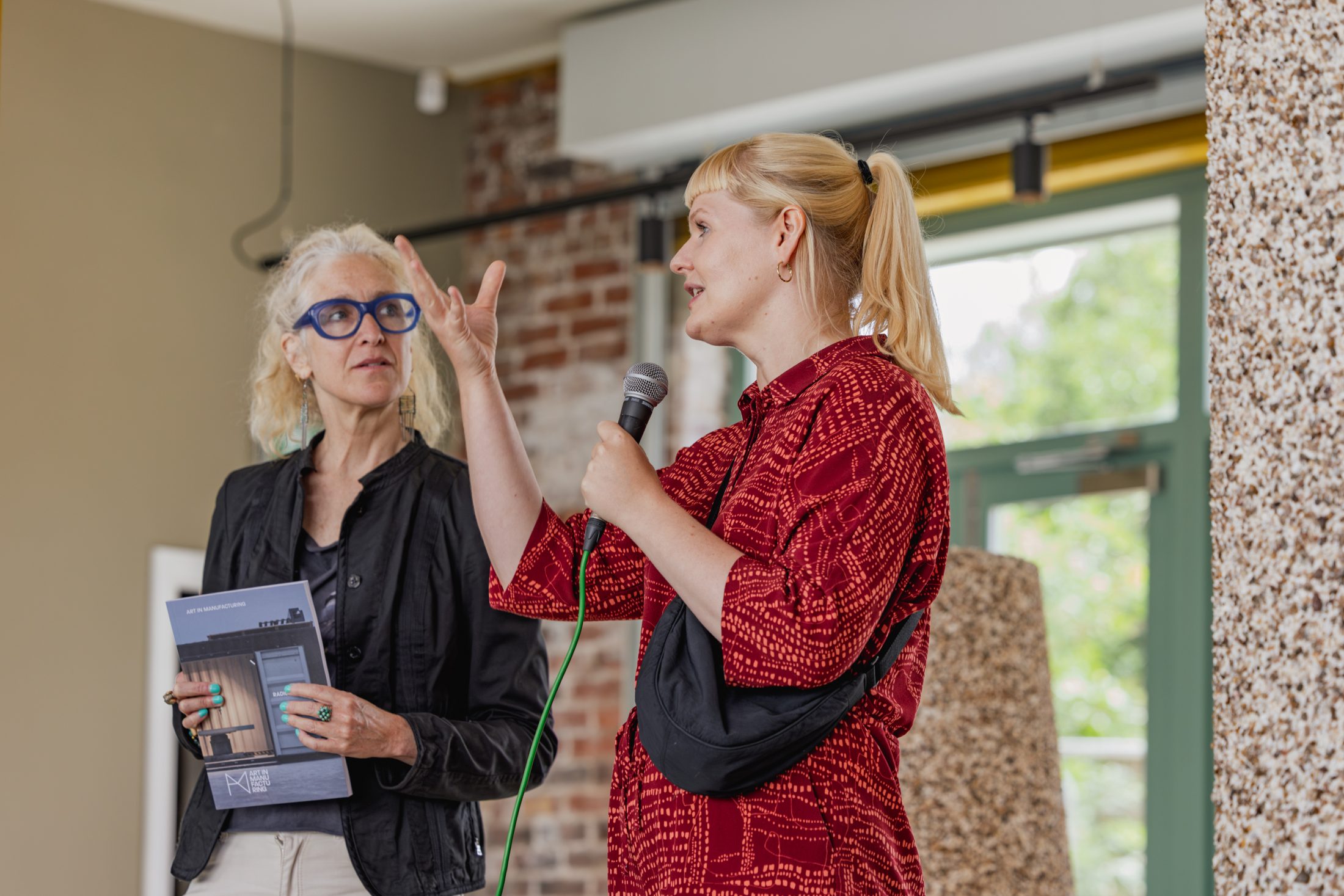 Two women indoors: one in a red dress holding a microphone speaks, gesturing with her hand; the other, in black with blue glasses, listens whilst holding a booklet. Brick wall and window are in the background.