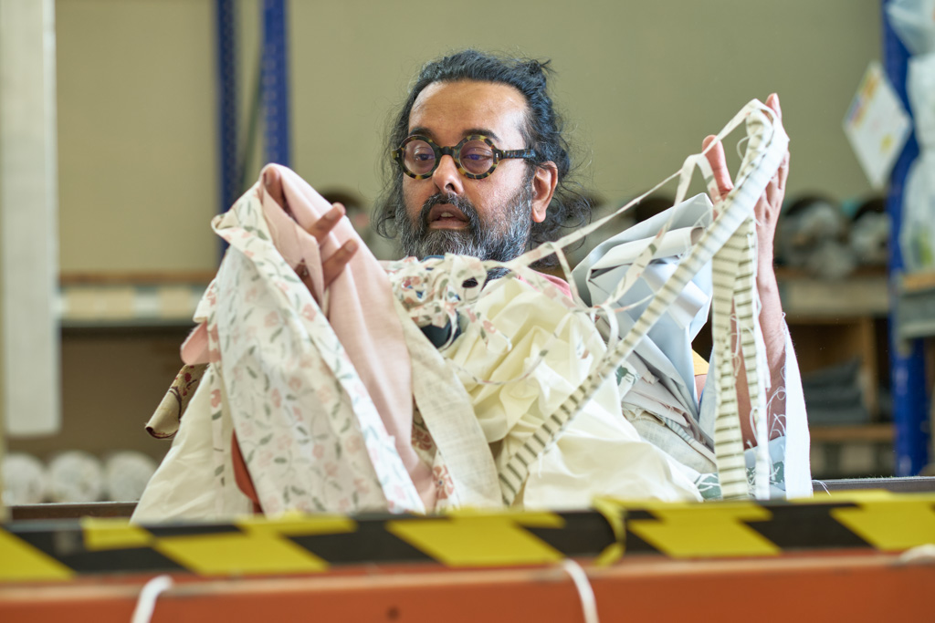 A person with glasses and a beard looks at a pile of assorted fabric pieces in an industrial setting, with caution tape on the foreground barrier.