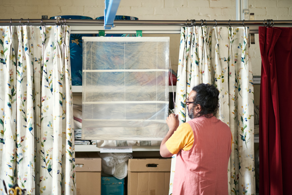 A person in a pink waistcoat and yellow shirt hangs a transparent storage organiser on a curtain rail, surrounded by floral curtains and boxes on shelves in the background.