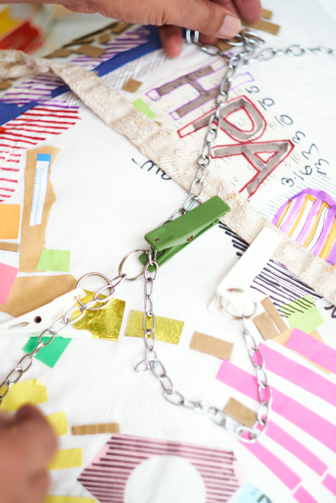 A hand adjusts a metal chain attached to a colourful, abstract collage with stripes, letters, and various paper pieces. The chain holds a green and a white clip, and HPA is visible among the artwork’s shapes and numbers.