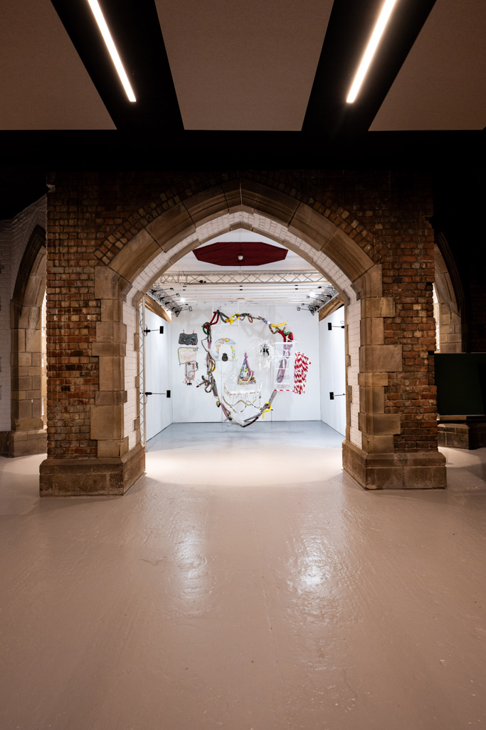An arched brick doorway frames a brightly lit art gallery room with a large abstract face mural on the far wall, surrounded by other colourful artworks. The space combines historic architecture with modern art displays.