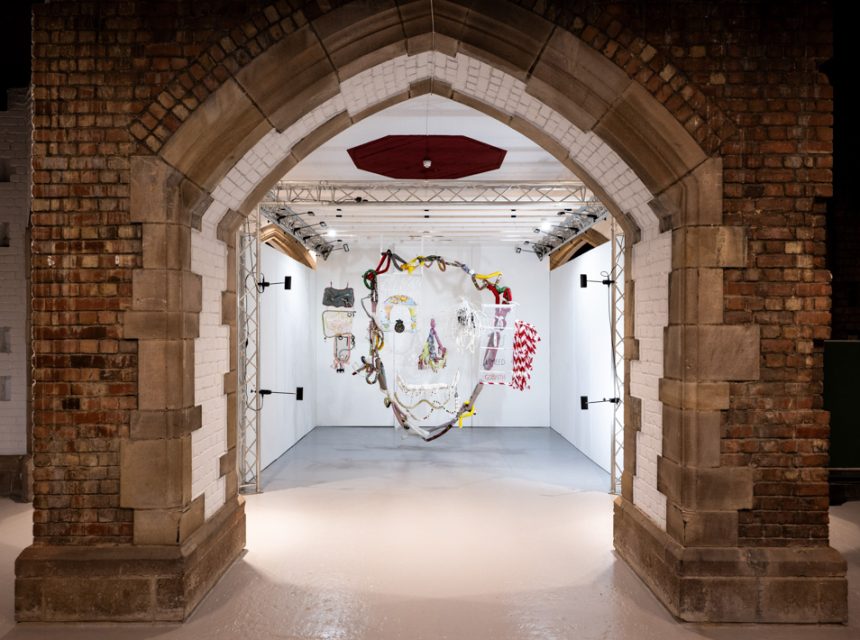 A white art gallery space is seen through a large stone archway with exposed brickwork. Colourful, mixed-media artwork is displayed on the back wall, surrounded by bright lighting.