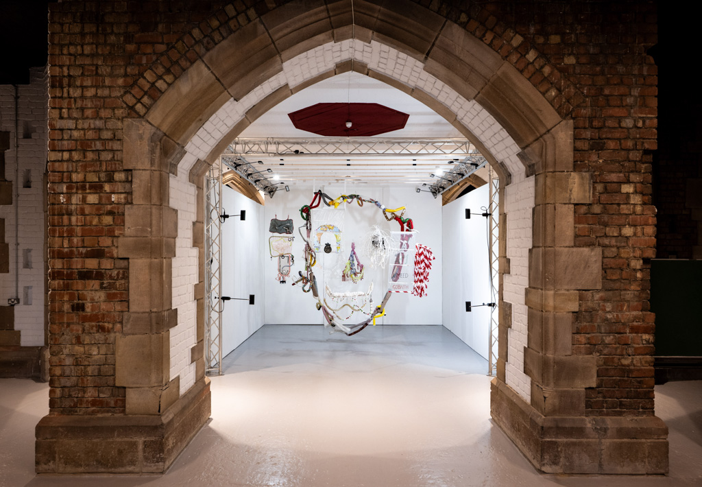A white art gallery space is seen through a large stone archway with exposed brickwork. Colourful, mixed-media artwork is displayed on the back wall, surrounded by bright lighting.