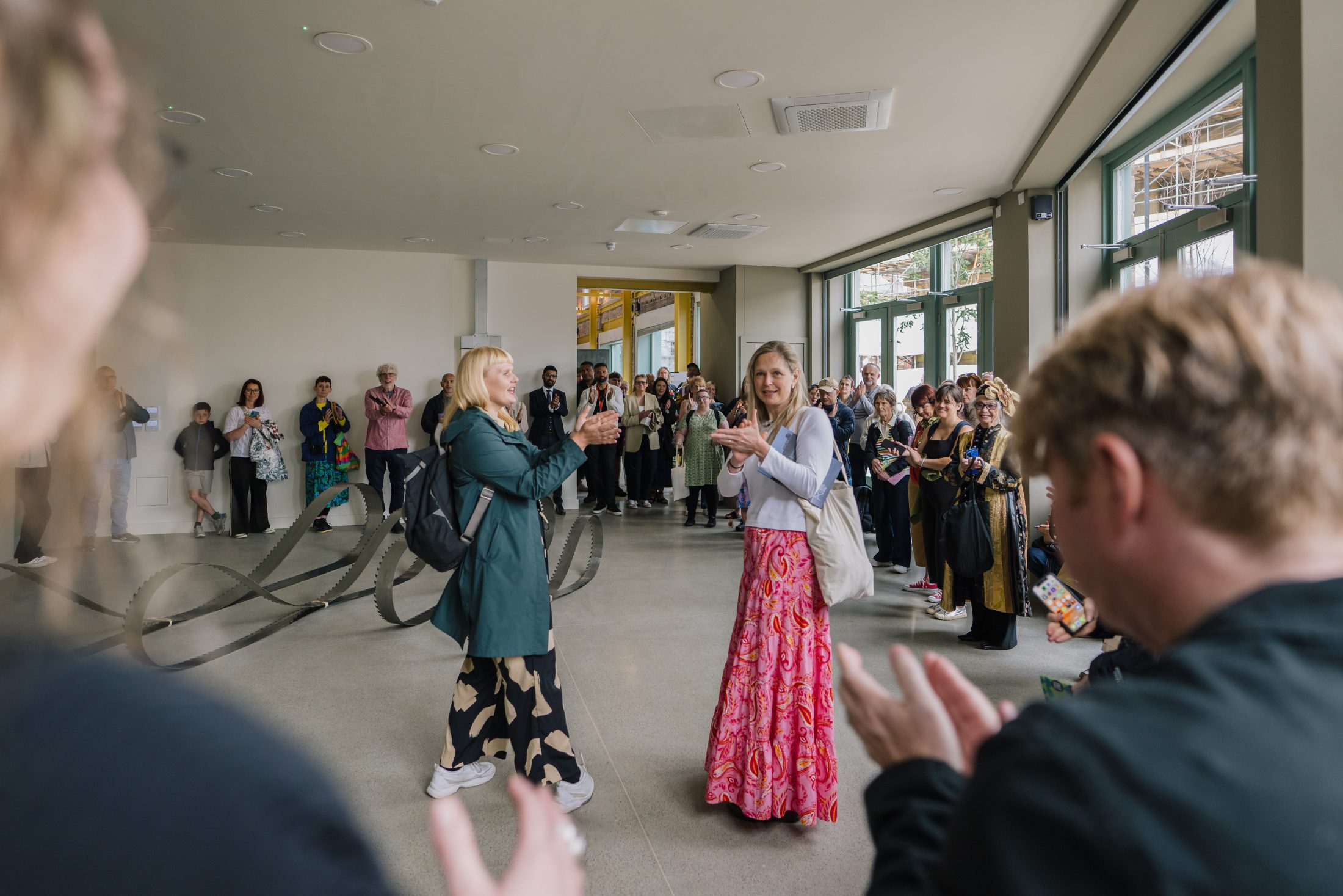 A woman in a green coat and patterned trousers stands and smiles as another woman in a white top and long pink skirt applauds her, surrounded by a crowd in a bright, modern gallery space.
