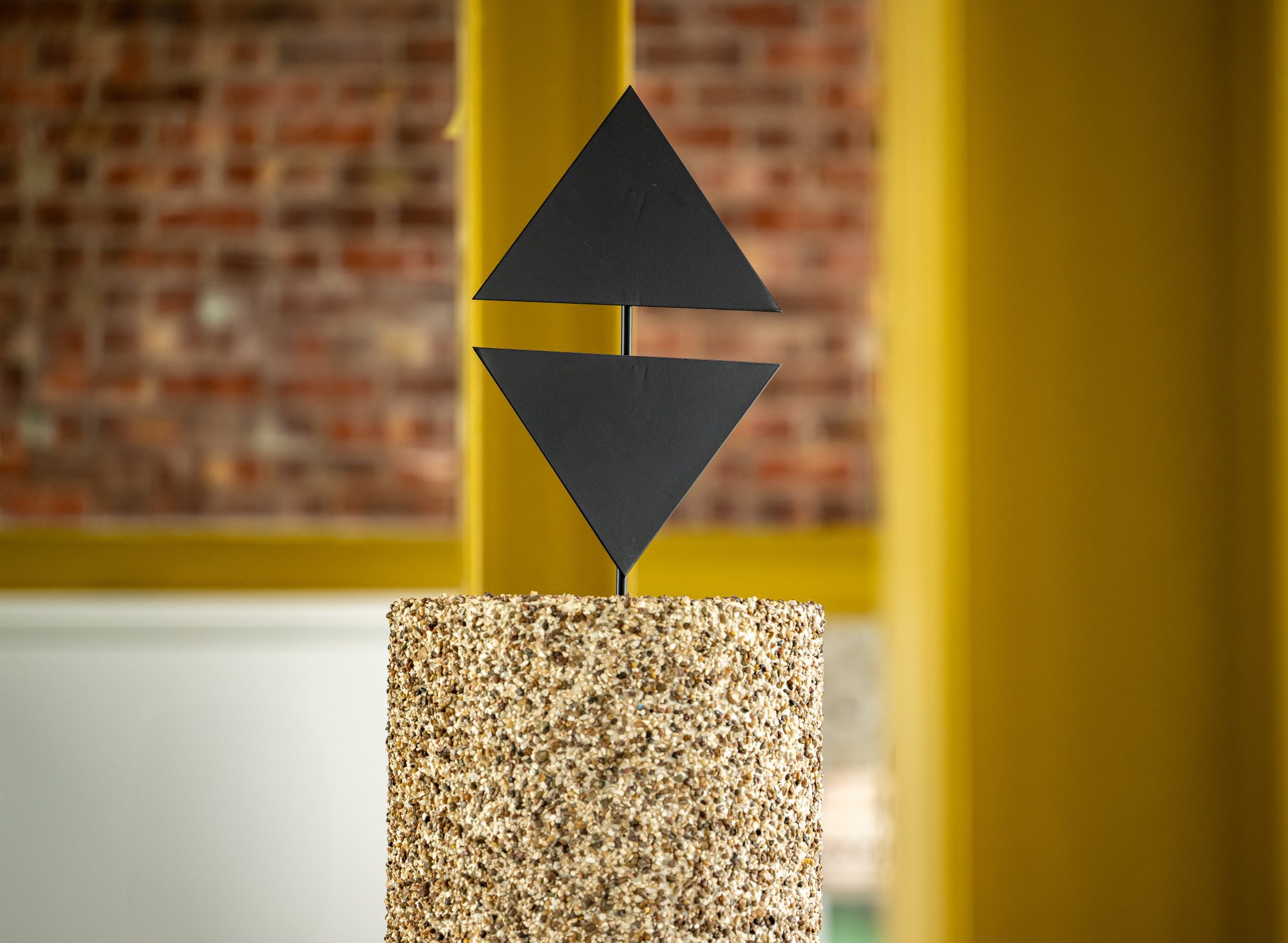 A sculpture featuring two black metal triangles, one pointing up and one down, separated by a gap and mounted on a cylindrical, textured stone plinth, with a blurred brick and yellow background.