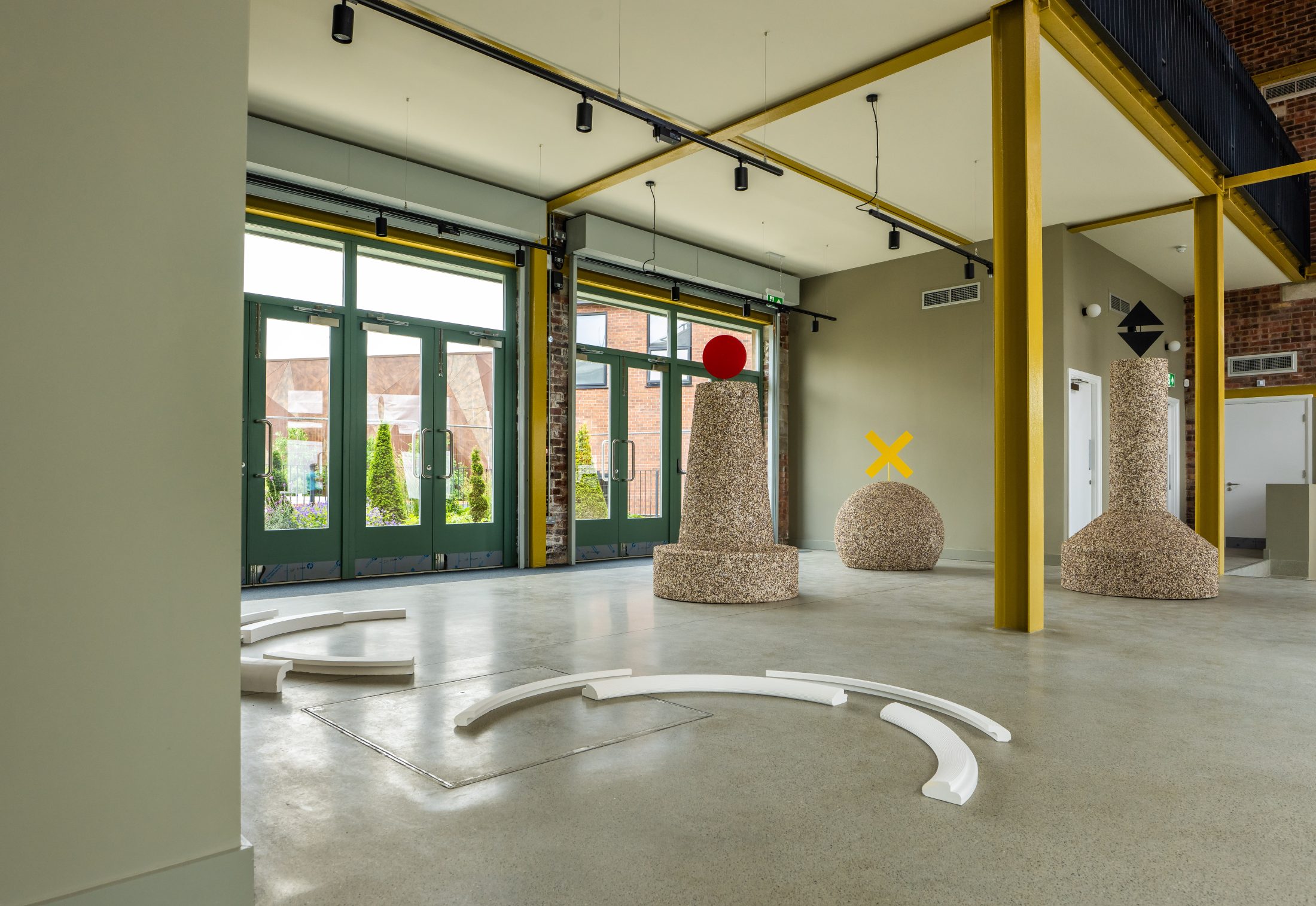 A modern art gallery with large windows, yellow columns, and abstract sculptures, including a tall stone structure with a red sphere, a round speckled sphere, and curved white pieces arranged on the polished floor.
