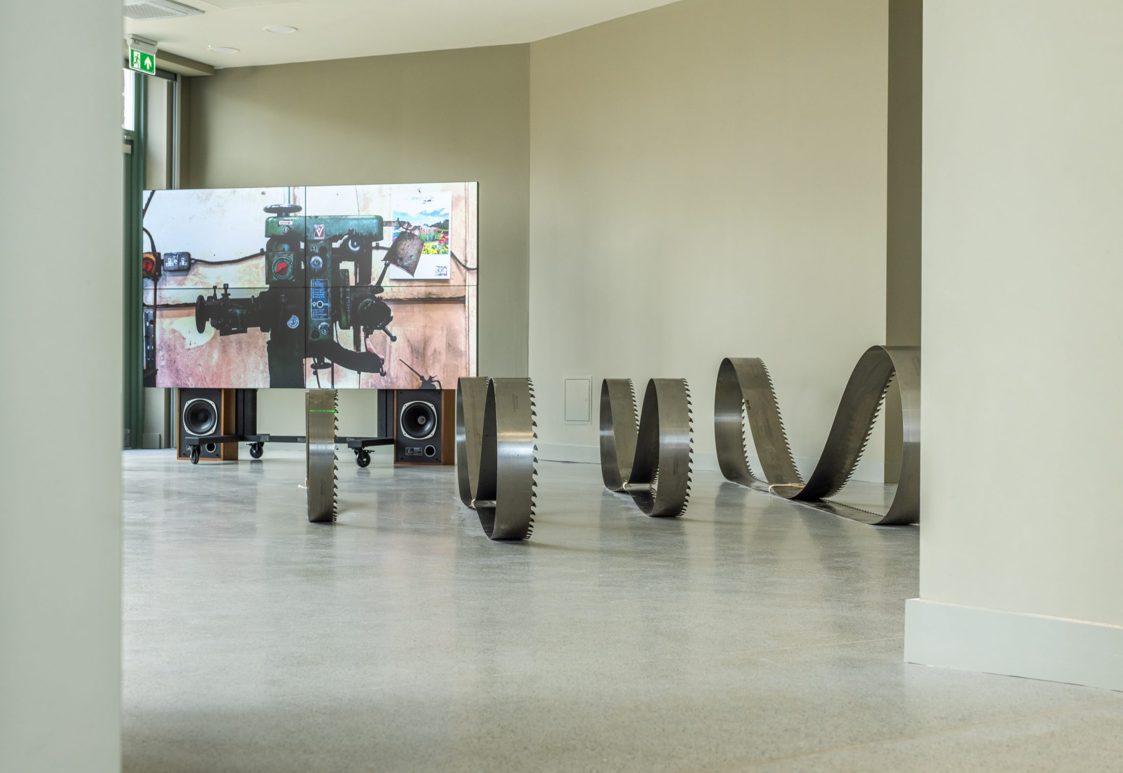 A modern art installation in a gallery features several large, metallic saw blades bent into wave shapes on the floor, placed in front of a video screen and two speakers against a beige wall.